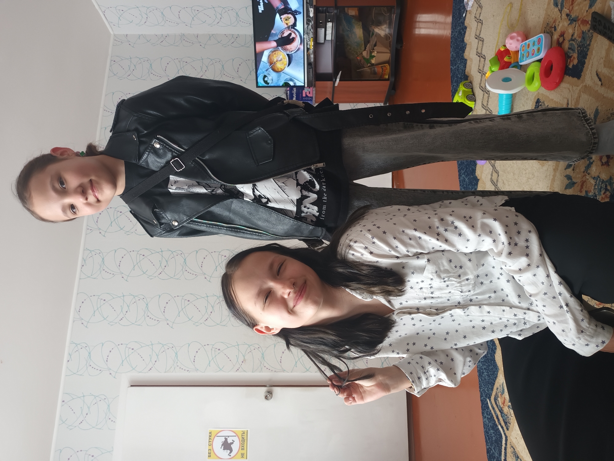This image depicts two young females posing in an indoor residential setting in Ольховка, Russia.
The subject on the right is a female standing, facing slightly left, looking towards the camera with a faint smile. She has dark hair pulled back and wears a black faux leather jacket over a dark t-shirt with a white graphic. A black crossbody strap extends from her right shoulder to her left hip. She wears dark grey jeans and a black belt with silver studs. A small green earring is visible in her left ear.
The subject on the left is a female kneeling on a patterned rug, facing the camera with her eyes closed and a wide smile. She has long dark hair, a nose ring, and is holding a strand of her hair between her left thumb and forefinger. She wears a white button-up shirt with a grey star pattern and dark pants.
The background features light-colored wallpaper with a repetitive pattern of intertwined light blue lines and small dots. To the left, a white door is visible, next to which a yellow rectangular sign with black text and a black bear icon reads "БЕЗ СТУКА НЕ ВХОДИТЬ!" (DO NOT ENTER WITHOUT KNOCKING!). To the right, a television set displays a cooking program and sits on a dark brown wooden cabinet. Various items, including boxes and bottles, are stored within the cabinet shelves, with some labeled "Nutrilak". The floor is covered by a patterned rug predominantly in blue, beige, and reddish-brown tones, with visible areas of an orange-brown floor underneath. Scattered on the rug in the foreground are several colorful children's toys, including a green toy car, a white and blue tube, a red ring, a green ring, a pink textured ball, and a white toy remote control with multi-colored buttons. A dark remote control is partially visible in the bottom right corner.