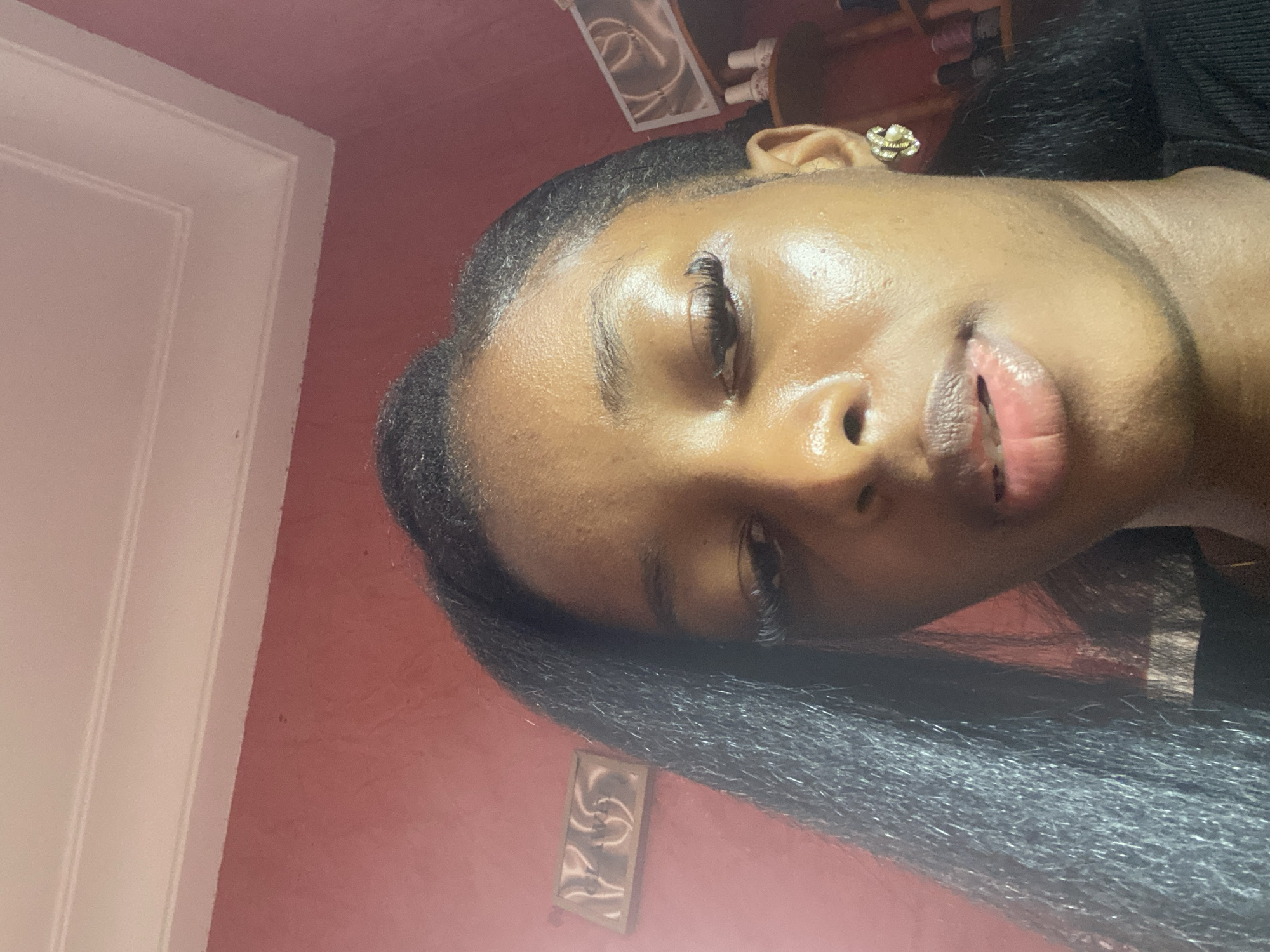 The image shows a close-up, upward-facing selfie of a young Black woman indoors. She has dark, straight hair, extended eyelashes, and is wearing a gold earring. Her skin is visible, showing some texture and shine. The background consists of a reddish-pink wall with decorative molding at the top. A framed picture is on the wall to the left, and shelving with various items, possibly cosmetics or craft supplies, is visible on the right. The lighting suggests an indoor setting, likely a home. The context provided states this scene is in the city of Jos, Nigeria, which implies the woman is a resident of or visiting Jos.