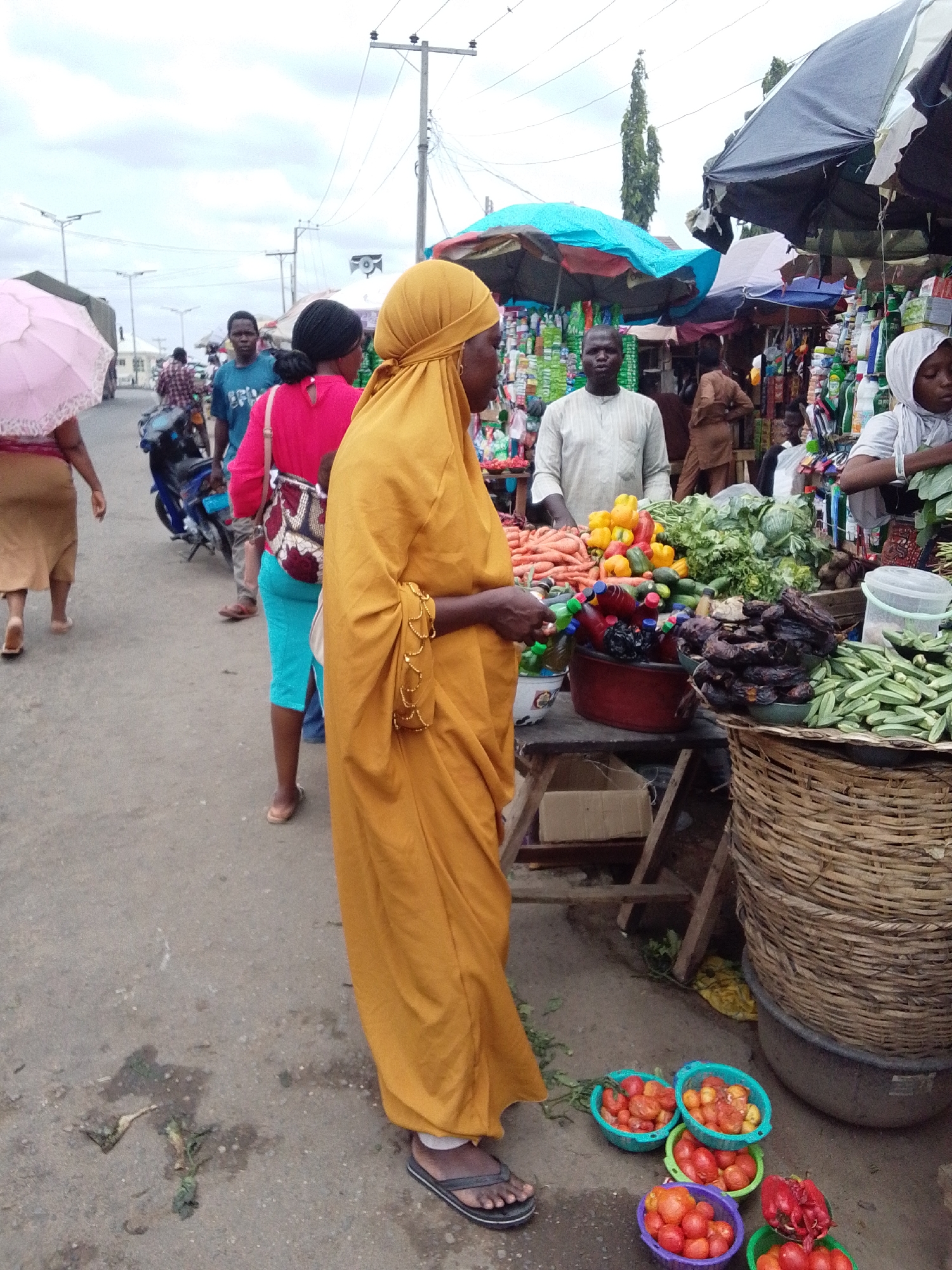 This medium shot captures a lively outdoor market scene in Lokoja, Nigeria, on an overcast day. The setting appears to be a street or open area dedicated to commerce.
In the immediate foreground, a woman stands with her back partially to the viewer, wearing a long, flowing mustard-yellow dress or abaya and a matching head covering. She wears black and white flip-flops. At her feet are several small, colorful plastic basins filled with fresh red tomatoes and some red bell peppers, suggesting she is either a vendor or a customer browsing.
To the right, a market stall is laden with a vibrant display of fresh produce. Visible items include piles of bright orange carrots, yellow and red bell peppers, green leafy vegetables like lettuce and cabbage, and numerous fresh okra pods spilling from a large, woven bamboo basket. Beside the okra, there's a heap of dark, dried items that appear to be dried fish or meat, and several plastic bottles containing beverages. Behind this stall, a man in a light-colored traditional tunic stands, looking towards the camera, seemingly a vendor. Another woman in a light headscarf is also visible behind the stall to the right.
Further into the scene, other individuals contribute to the bustling atmosphere. On the left, a person holds a pink umbrella, walking away from the camera. Another woman in a bright pink top and turquoise capri pants walks towards the left, carrying a patterned bag. Motorcycles are parked or moving in the background, and utility poles with power lines and streetlights frame the sky. The paved ground shows scattered vegetable scraps and general market debris, characteristic of an active marketplace. The overall impression is one of daily commerce and community activity. No legible text is visible in the image.