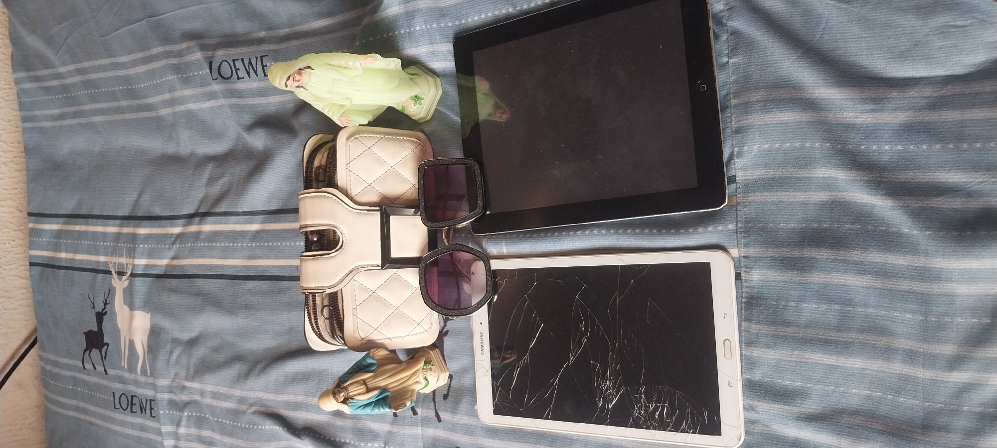 The image displays a flat lay of personal items on a blue and white striped fabric, likely a bedsheet or blanket. The items are arranged on a bed, which suggests a domestic setting. The lighting is natural and diffused, indicating it might be daytime, possibly indoors near a window.
The main subjects are two tablet devices, a white quilted clutch bag, and a pair of sunglasses. One tablet is black and appears to be an Apple iPad. The second tablet is white and has a visibly cracked screen; it appears to be a Samsung tablet. The clutch bag is open, revealing some contents that look like money and cards. The sunglasses have large, dark lenses and a chunky frame.
Also present are two small figurines. One is a pale green, glowing figurine of a robed person, possibly religious. The other is a small figurine of the Virgin Mary, depicted with a blue robe. The fabric features a repeating pattern of horizontal stripes in varying shades of blue and white, with some thin black stripes interspersed. On the left side of the fabric, there are silhouettes of deer. Visible text includes "LOEWE" printed in dark blue letters on the fabric, near the top left and bottom left corners. The scene is static, with no discernible activity taking place; the items are simply laid out. The overall impression is one of everyday personal belongings.