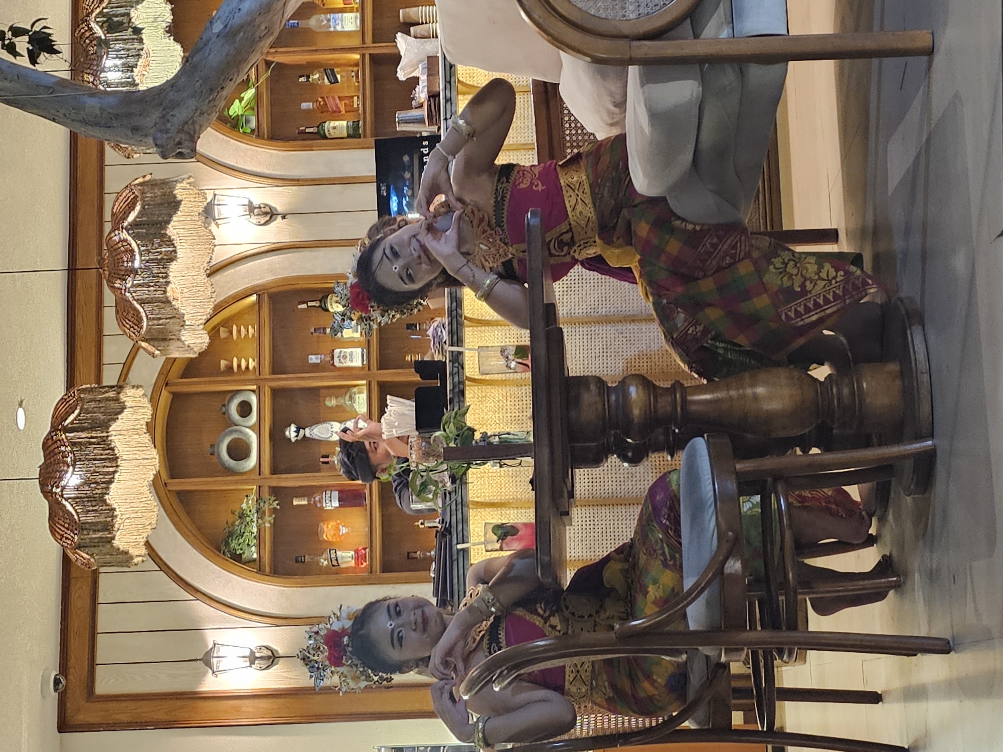 The photograph depicts two Balinese women, dressed in traditional attire, positioned at separate tables within a restaurant. Both women are wearing elaborate headpieces and traditional Balinese outfits consisting of sarongs and tops, with intricate gold jewelry. They appear to be either posing for a photograph or in the midst of a cultural performance or presentation.
The background reveals a restaurant interior. There's shelving stocked with an array of liquor bottles and decorative items. The lighting is warm, provided by ornate lamps with textured shades. Additional tables and chairs are visible, with neutral-colored upholstered seating.
The restaurant is located in the city of Denpasar, Indonesia.