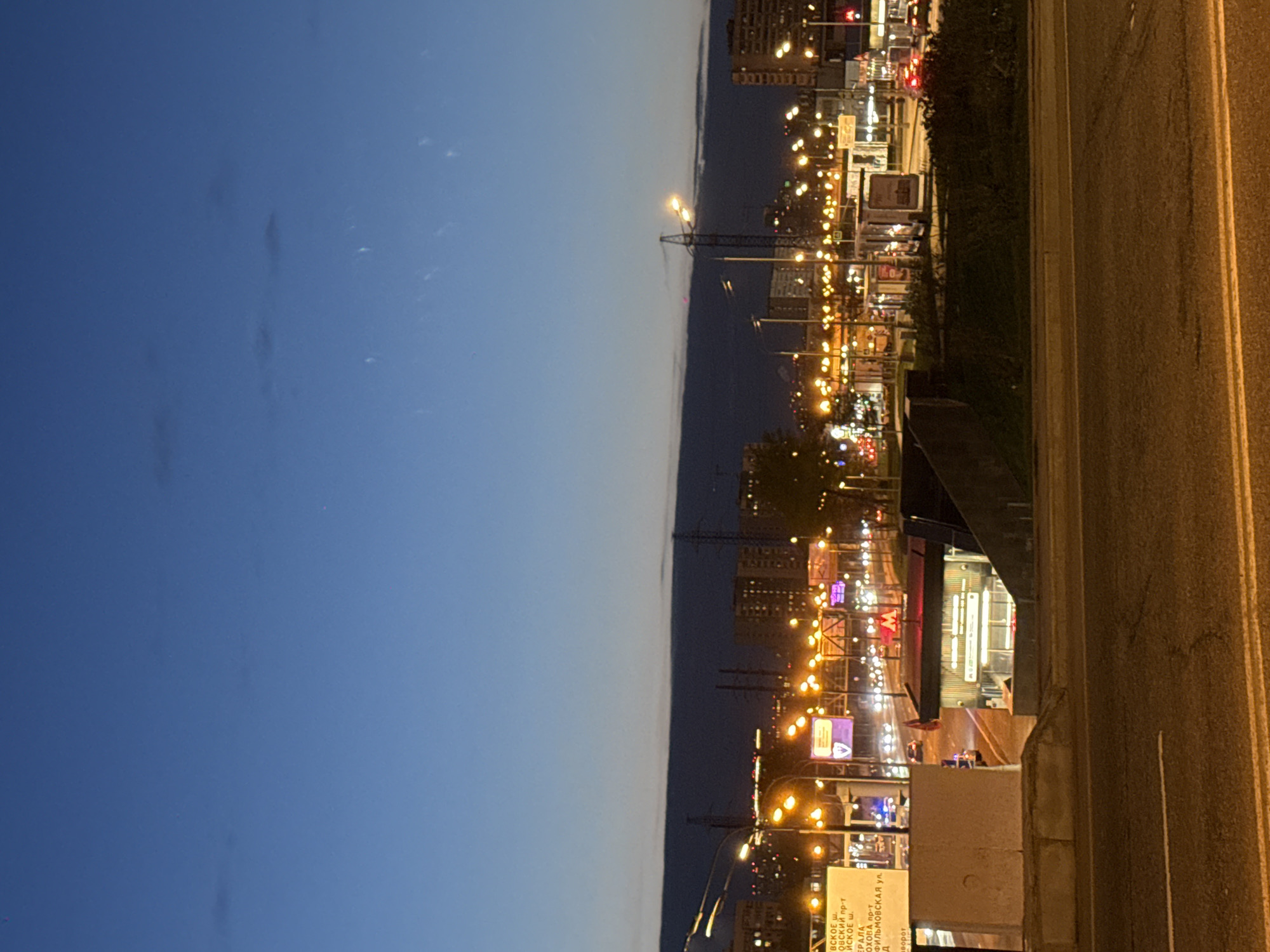 The image, rotated 90 degrees, captures an urban landscape at twilight or dusk, likely in Khimki, Russia. A deep blue sky with faint, wispy clouds dominates the upper half. Below, a city comes alive with numerous warm-toned streetlights and illuminated buildings, creating a vibrant glow against the fading light.
On the right side, a multi-lane asphalt road with clear yellow double lines is visible. To the left of the road, various structures and elements compose the cityscape. A prominent tall, dark structure, possibly a crane or power line tower, is silhouetted against the horizon. Below the road level, an entrance featuring a distinctive red "M" sign, characteristic of the Moscow Metro, suggests a subway station or underpass.
Several billboards and street signs with Cyrillic text are visible. OCR analysis reveals fragments of street names such as "ФИЛЬМОВСКАЯ ул." (Filmovskaya ulitsa - Filmovskaya Street), and parts of others ending in "ШОССЕ" (Shosse - highway) and "ПРОСПЕКТ" (Prospekt - avenue). Other readable text includes "FIT" (likely a business name) and a partial word "ворог" (possibly part of "поворот" - turn). The illumination implies ongoing city life and activity, transitioning from day to night.