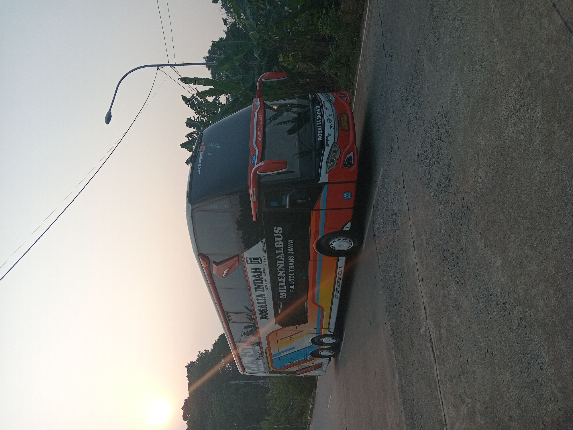 A double-decker bus, branded "ROSALIA INDAH MILLENNIALBUS", is parked on a paved road. The bus is predominantly red and white with colorful graphics along its side. The sky is clear and bright, with the sun visible in the upper left corner, casting a warm glow. Green trees and banana plants line the right side of the road, and a street light is visible in the background. The scene appears to be a roadside in a semi-urban or rural area, likely Depok, Indonesia, given the context provided.