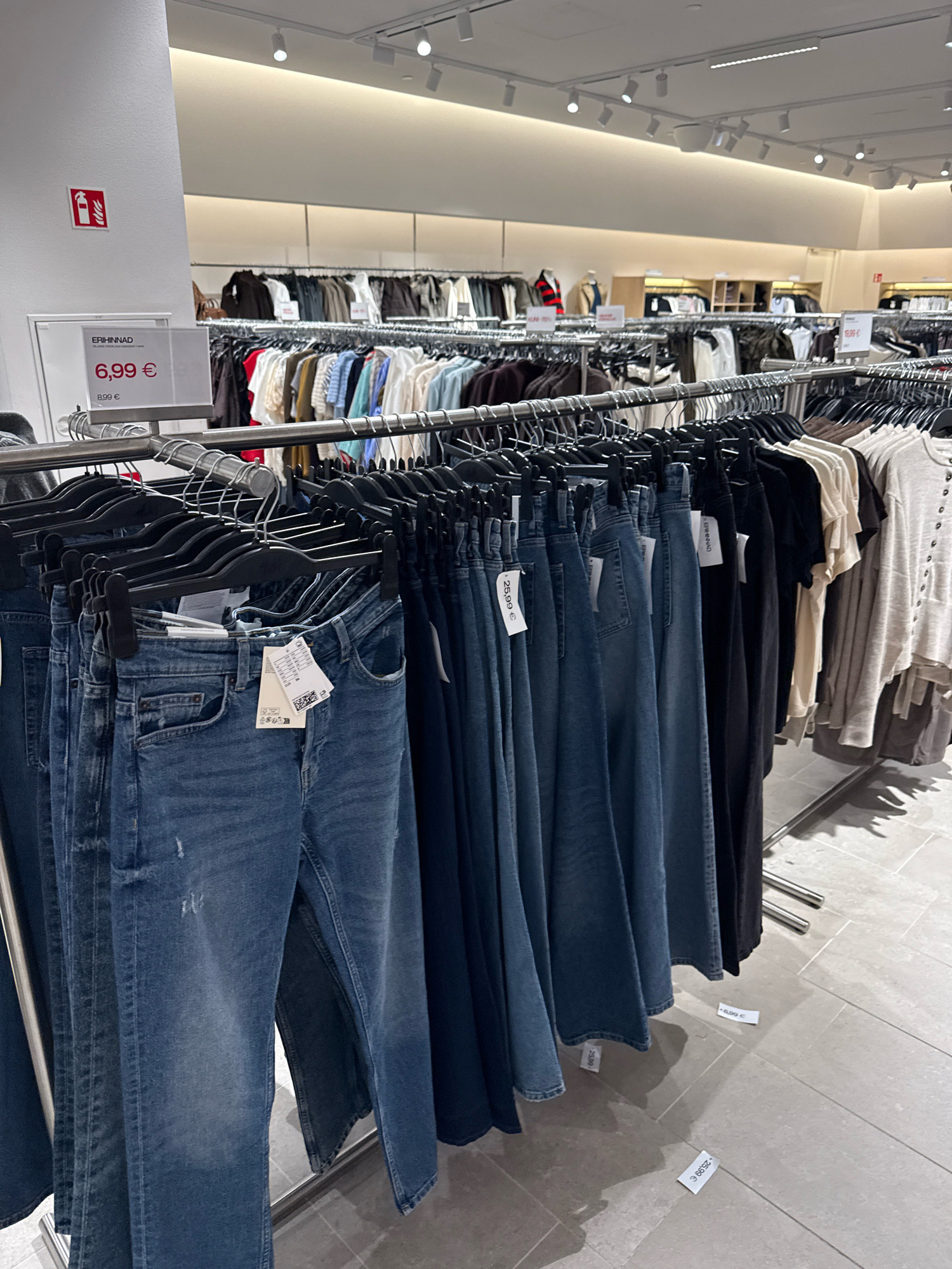 The image shows a clothing store interior, likely a department store, with racks of clothing and signage. A price tag on a white sign reads "ERI HINNAD" and "6,99 €", indicating special prices. Below this, another price of "8,99 €" is visible. The clothing on display includes denim jeans, t-shirts, and other apparel. There are price tags on some of the clothing items, with one visible price tag reading "25,99 €". The floor is tiled. In the background, more clothing racks and store fixtures are visible. The overall impression is a retail environment focused on clothing sales. The presence of the Estonian language on the price tag ("ERI HINNAD") strongly suggests that the image was taken in Estonia. Given the context of a retail store with prices in Euros and signage in Estonian, it is highly probable that this scene is located in Tallinn, the capital city of Estonia.