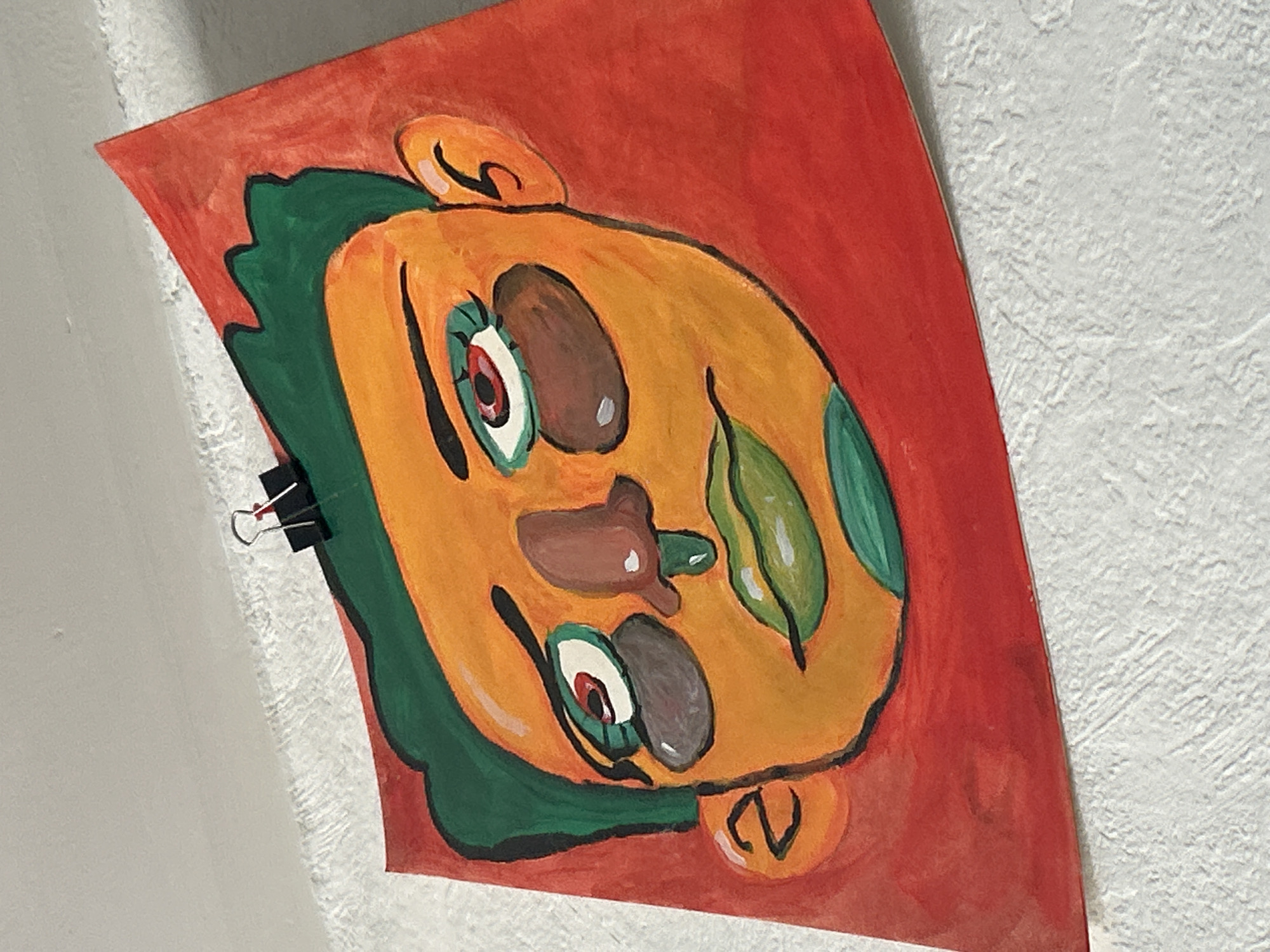 The image depicts a colorful painting of a face on a red background, attached to a white textured wall with a black binder clip. The face is predominantly yellow and orange, with green hair and green leaf-like shapes on the face. The eyes are outlined in green with red pupils. The nose is brown. There are small, orange ears on both sides of the head. The location is an interior setting, the exact city and place is Unknown.
