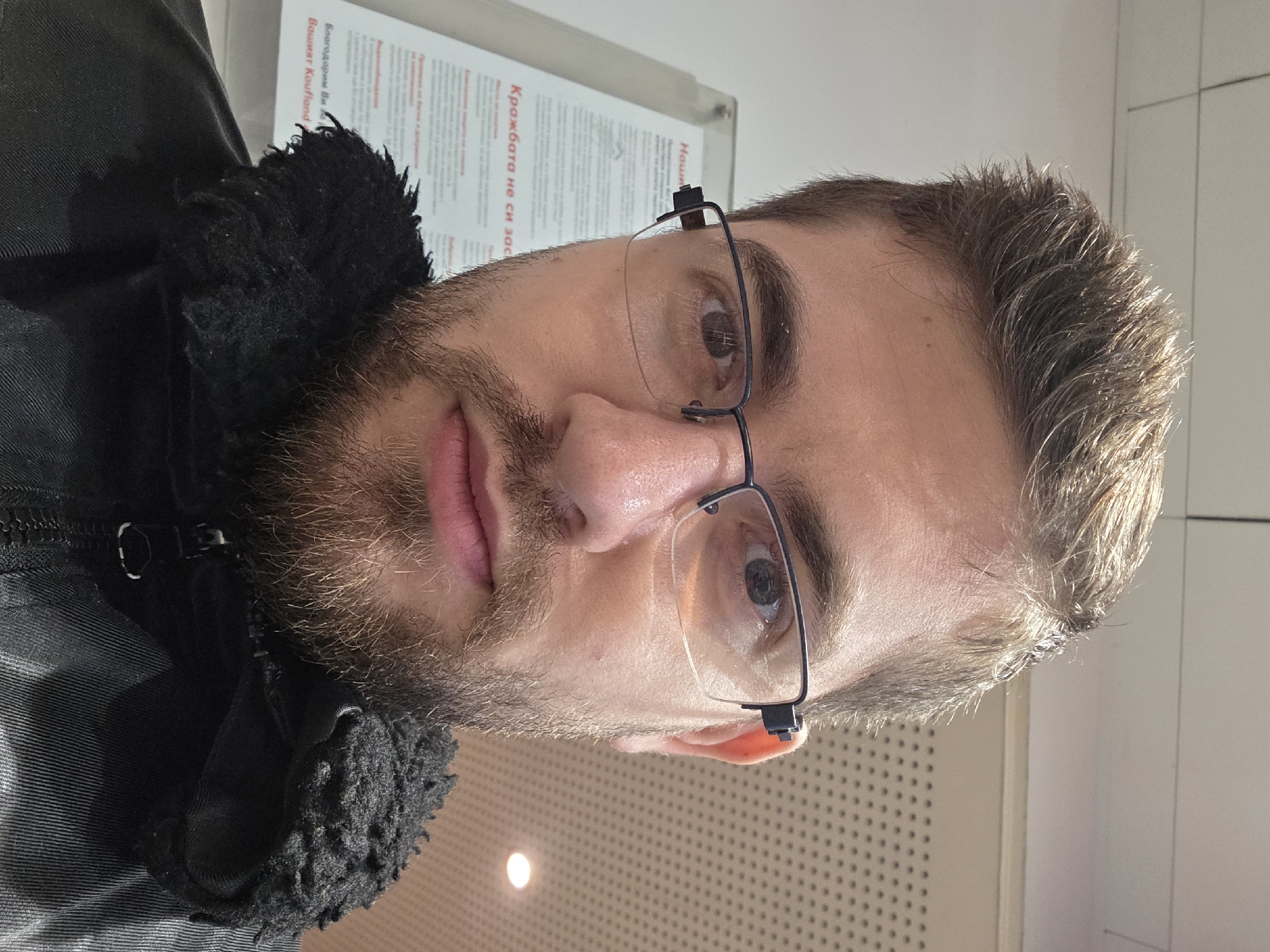 The image shows a close-up selfie of a Caucasian man with short brown hair, a beard, and glasses. He is wearing a black jacket with a fur-like collar. In the background, out of focus, is a poster with text in Bulgarian. Given the presence of Bulgarian text, it is plausible the image was taken in Plovdiv, Bulgaria, but this cannot be confirmed from the image alone. The man's expression is neutral. The environment appears to be indoors, possibly a public space.