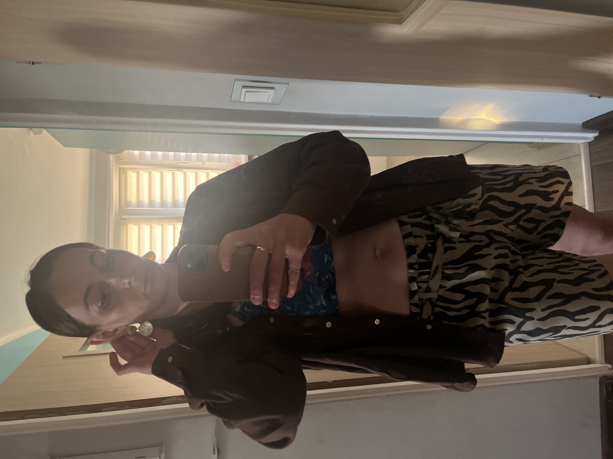 A woman is captured in a mirror selfie in what appears to be a hallway or entryway. She is wearing a brown open button-down shirt over a floral crop top, revealing her midriff. She also has on animal-print shorts. She is adjusting one of her large, dangling earrings. The background shows a doorway, a light switch on the wall, and a window with blinds. The lighting is natural, coming from the right side of the frame, casting a warm glow.