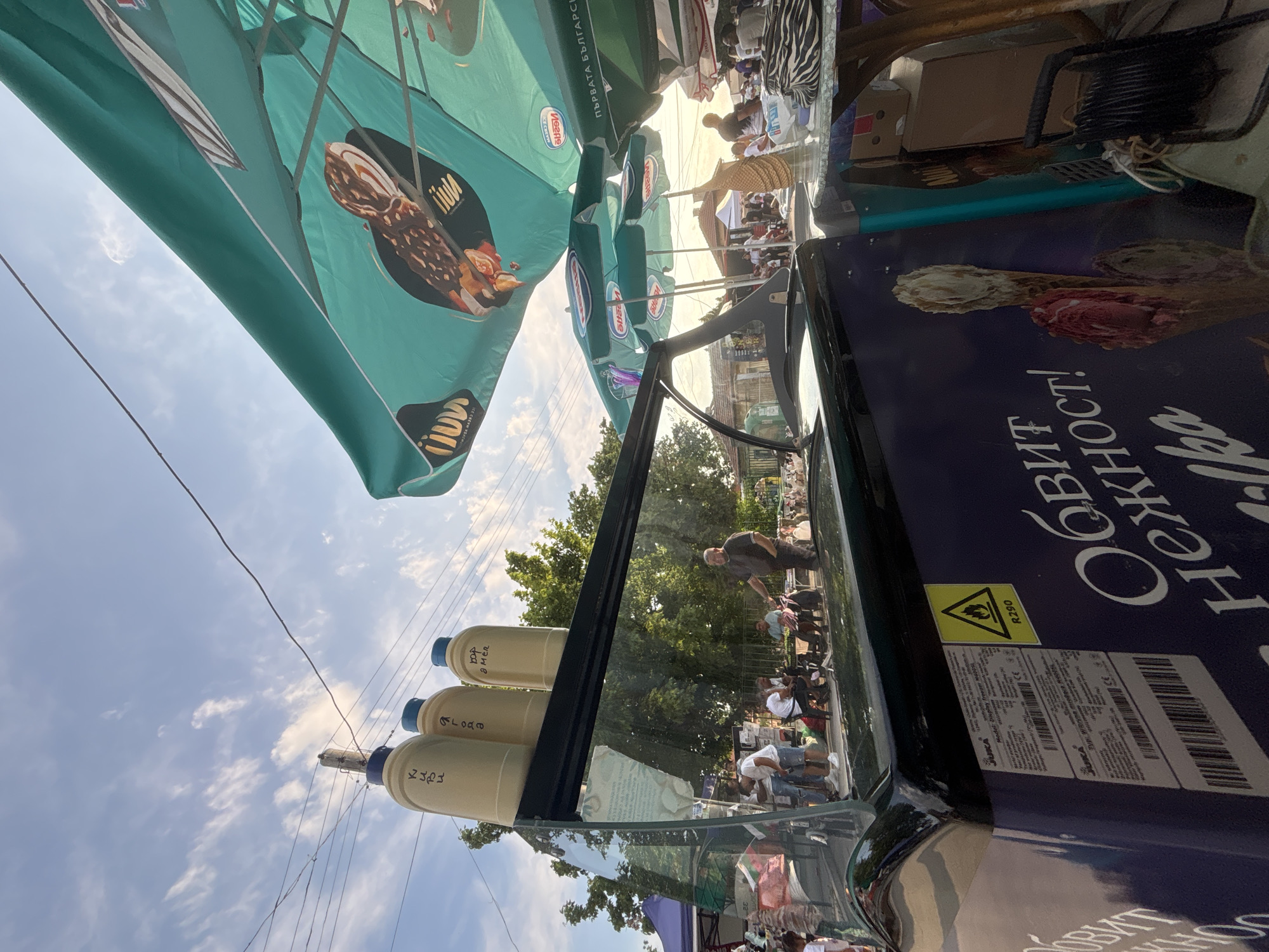 This detailed description outlines a vibrant outdoor market scene in Kostievo, Bulgaria, captured during daytime under a partly cloudy sky.
In the immediate foreground, a dark blue/purple ice cream display freezer is positioned at an angle. Its side features large white Cyrillic text, "ОБВИТ НЕЖНОСТ!" (Embrace Tenderness!) and "ilka", accompanied by a graphic of an ice cream cone containing vanilla and red scoops. A yellow triangular warning label with a flame symbol and "R290" is affixed to the freezer. A technical label lists "JUKA", "Model: Display Freezer H40SL", and other specifications.
On top of the freezer's glass display, three oversized, light-colored decorative bottles with blue caps are arranged. The leftmost bottle displays Cyrillic text "КЪПИ" (KAPI), the middle one "ЯГОДА" (YAGODA - Strawberry), and the rightmost "ВОР АМЕН" (VOR AMEN). The reflective glass surface of the freezer distorts the background elements.
Behind the freezer, a bustling area is visible. A man in a dark shirt and trousers stands near a small table, interacting with a group of casually dressed individuals, some seated and some standing. To the right of the freezer, a table holds a woven, pineapple-shaped decorative object and a black and white zebra-print bag.
Multiple large, teal-colored market umbrellas provide shade across the scene. The umbrella directly above the freezer is branded with a reversed "Nestlé" logo and an image of an ice cream bar, along with the Cyrillic text "ПЪРВАТА БЪЛГАРСКА" (THE FIRST BULGARIAN) and reversed "ICE CREAM YCHACH". Other similar "Nestlé" branded umbrellas are visible further back.
The upper portion of the image is dominated by a bright blue sky with scattered white cumulus clouds. Electrical wires crisscross the sky, supported by a utility pole. Green deciduous trees line the background, providing a natural backdrop to the market. In the distant background, low-rise buildings with tiled roofs, characteristic of the region, are faintly visible, and more people are dispersed throughout the area, indicating a lively community gathering or market event.