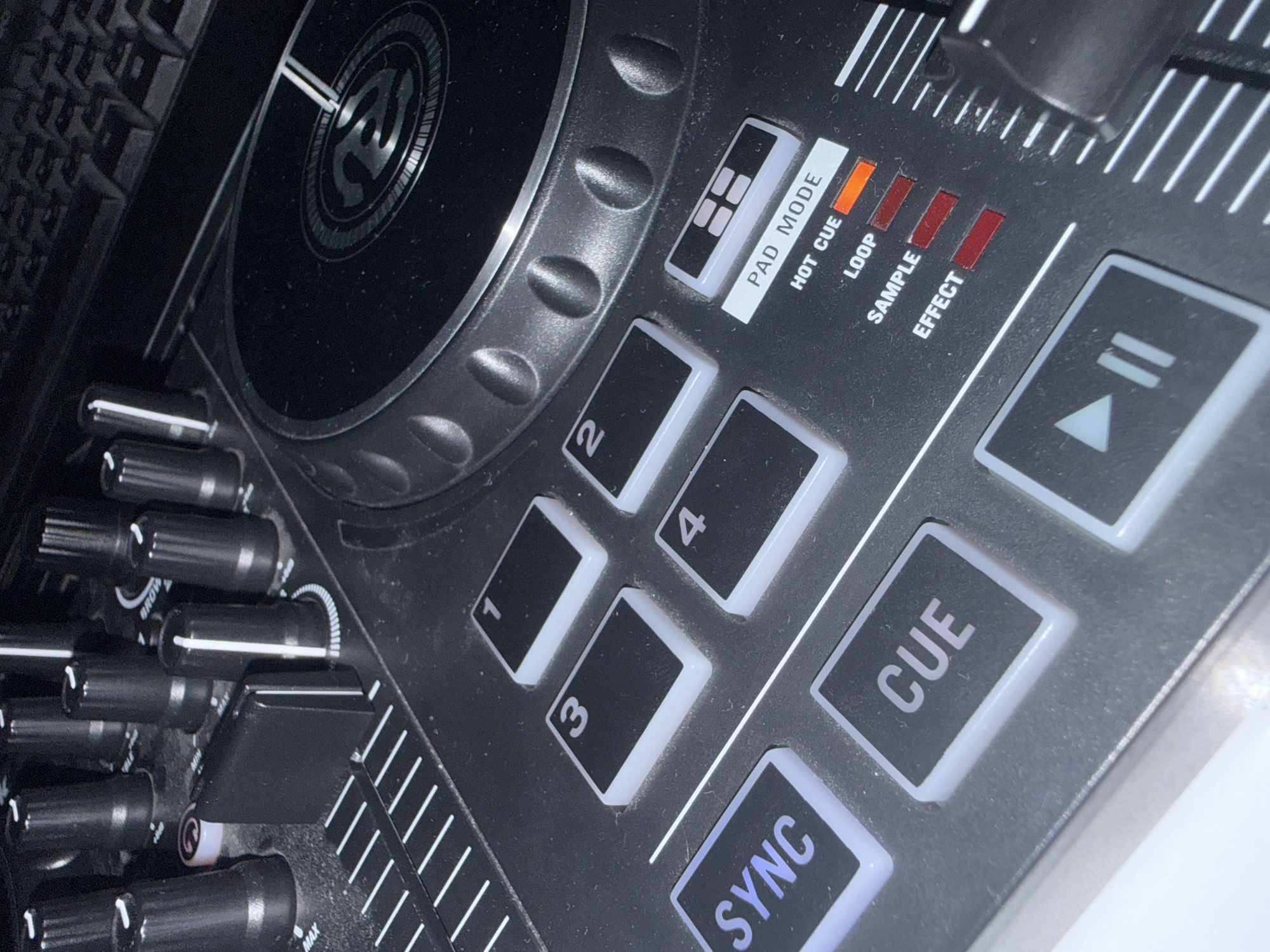 This is a close-up, eye-level, indoor shot of a black DJ controller. The controller is angled from the left foreground to the right background, with a portion of a computer keyboard visible in the upper right corner. The DJ controller features several illuminated buttons and indicators. Notably, there are four illuminated square buttons labeled "1", "2", "3", and "4", and two larger illuminated buttons labeled "SYNC" and "CUE". Below the "PAD MODE" button, there are four vertical LED indicators, with the top one labeled "HOT CUE" illuminated in orange, and the other three labeled "LOOP", "SAMPLE", and "EFFECT" showing red illumination. Several black knobs with white indicator lines are visible in the upper portion of the image, along with a textured circular jog wheel. The overall surface of the controller appears dusty. The scene's location in Burgas, Bulgaria, cannot be determined from the provided image.
