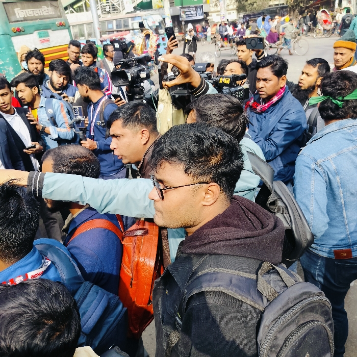 A crowd of people fills the frame, many holding cameras and phones. Several appear to be journalists or media personnel, indicated by professional video equipment. Individuals wear a mix of casual clothing, including jackets, jeans, and backpacks. One person wears glasses and a hoodie, facing sideways. A bus with writing on the side is partially visible in the background. Others wear jackets and light sweaters.
