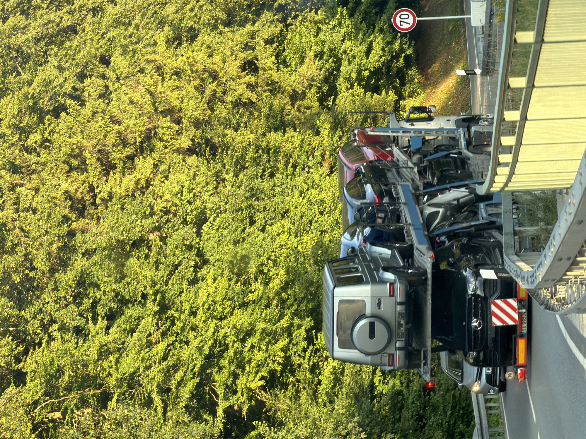 The image depicts a car transporter truck carrying several vehicles on a road. The truck is a commercial vehicle with a multi-level trailer loaded with cars. The top level of the trailer has a silver SUV, and below it are other cars, including a black Mercedes-Benz SUV in the lower section. The truck is in motion, indicated by the blurred background and the speed limit sign of 70 visible on the right side of the road. The environment is characterized by a lush green treeline on the left, suggesting a rural or suburban setting. The road appears to be a highway or a major road with guardrails. The image is taken from a slightly elevated perspective, looking towards the front of the truck. There are no people clearly visible in the image. The scene appears to be captured during daylight, with sunlight illuminating parts of the foliage and the road. The location is specified as Grainau, Germany.