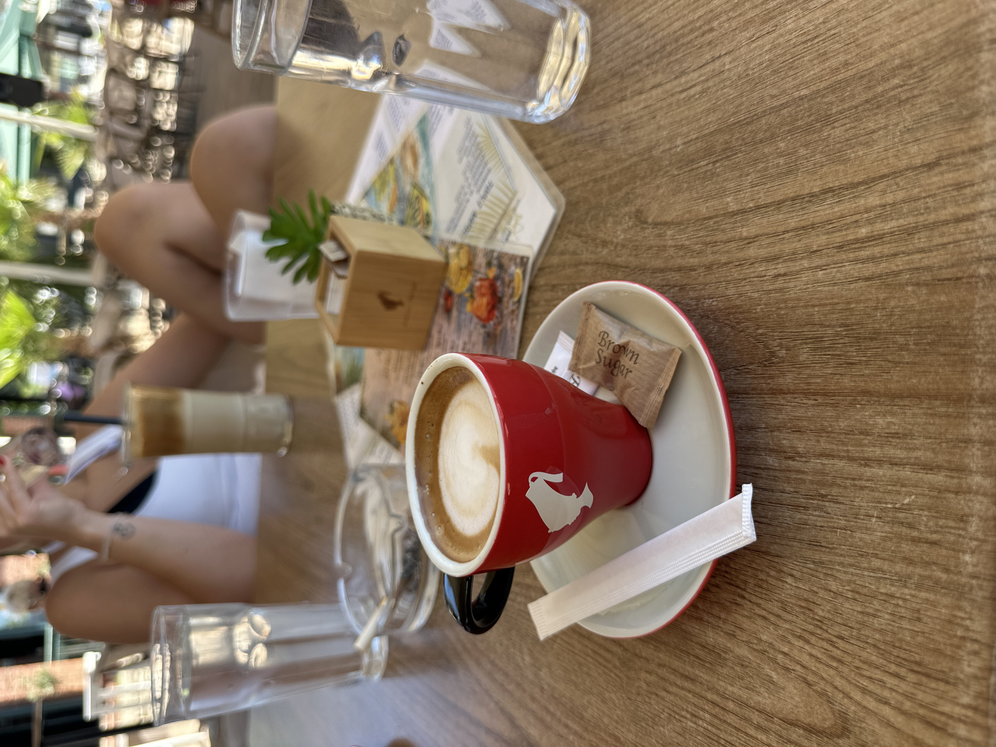 The image shows a close-up of a red coffee cup with a white saucer and a brown sugar packet on a wooden table. A white stirrer is placed next to the cup. In the background, there are glasses of water, an ashtray with a cigarette, and a person is visible, partially out of focus, with trees and outdoor seating in the background. The overall scene suggests a casual outdoor cafe setting. The location is specified as Boreti, Montenegro.