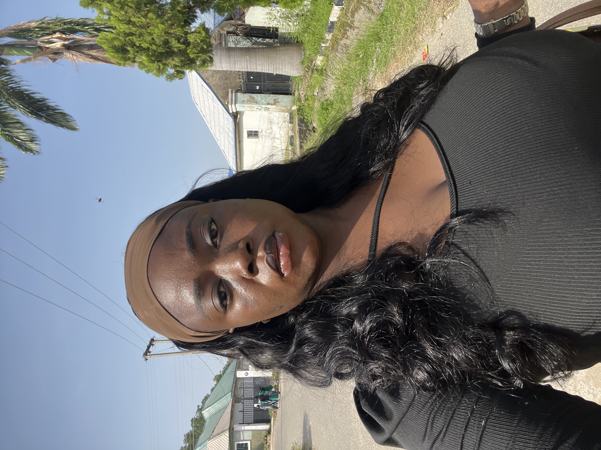 The image is a selfie of a woman wearing a brown headband and a black, ribbed top. She has dark, wavy hair that falls over her shoulder. Her skin appears shiny, possibly due to perspiration or oil. She is wearing a silver watch on her wrist.
In the background, there is a clear blue sky, power lines, palm trees, and buildings. One building has a white roof with a gabled design. Another building has a green roof. There is a road and some grass visible. The image appears to be taken outside during the day.
Based on the architecture and vegetation, it is likely the photo was taken in a residential area in Abuja, Nigeria.