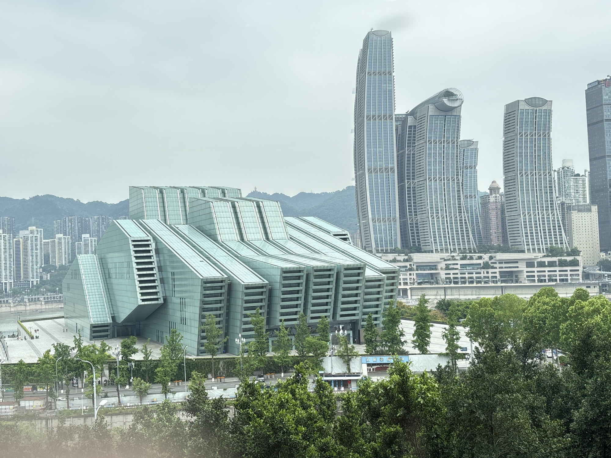 This wide-angle shot shows 江北城街道, China, with a foreground of green trees partially obscuring a street lined with trees and streetlights. Behind the trees is a striking modern building composed of tiered, glass-covered structures. To the right of the building, several skyscrapers dominate the skyline. In the background, more distant mountains and buildings are visible under an overcast sky. A large paved area is located in front of the modern building. People can be seen walking in the distance on the pavement.