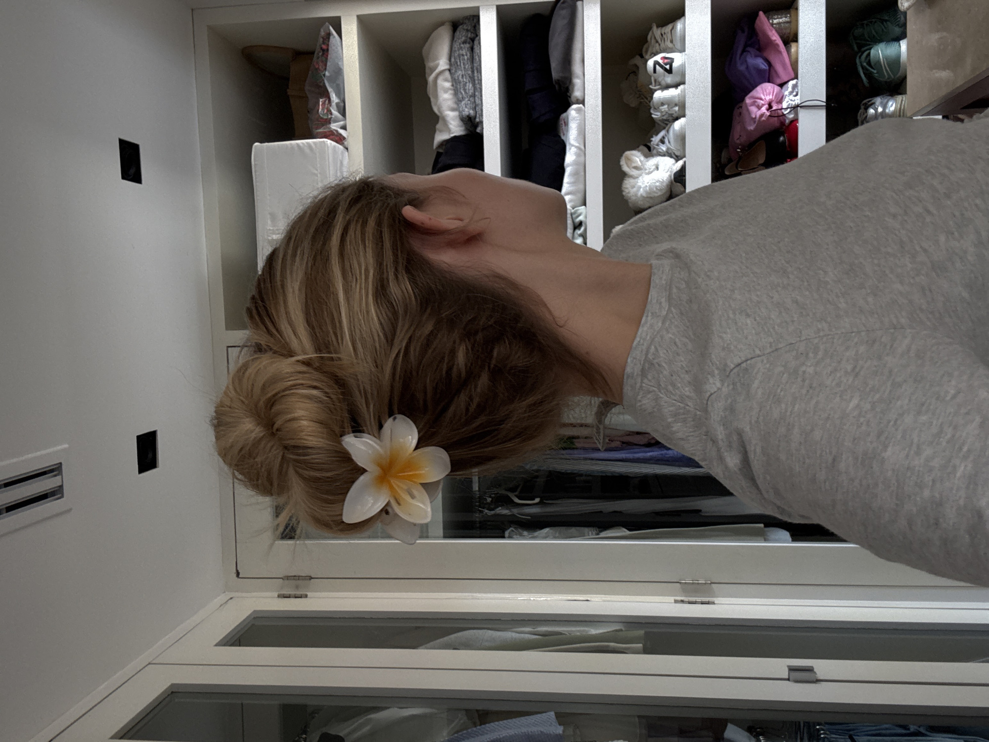 A woman in a gray top is near a white closet. The woman has her hair in a bun with a white and yellow flower. The closet has shelves with clothes and shoes. The location is not discernible from the image.