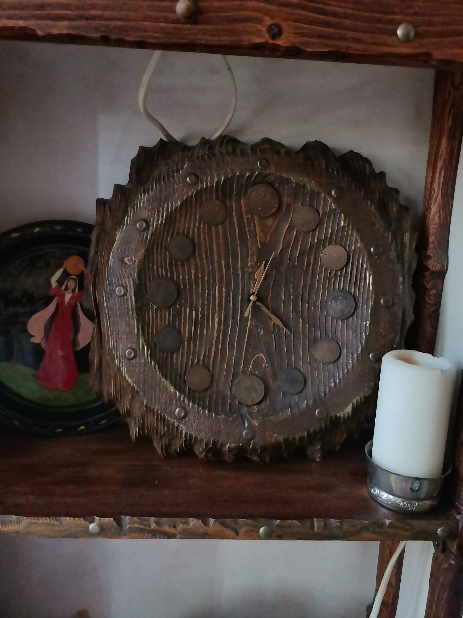 The image shows a wooden clock on a shelf. The clock has a rough, textured surface and is decorated with what appear to be coins. There is also a decorative plate with a woman in a red dress holding a jug, and a white candle in a silver holder on the shelf. The shelf itself appears to be made of dark wood and is part of a larger wooden structure, possibly a bookcase or cabinet. The background is a plain, light-colored wall. The overall impression is of a rustic or handcrafted decorative display.