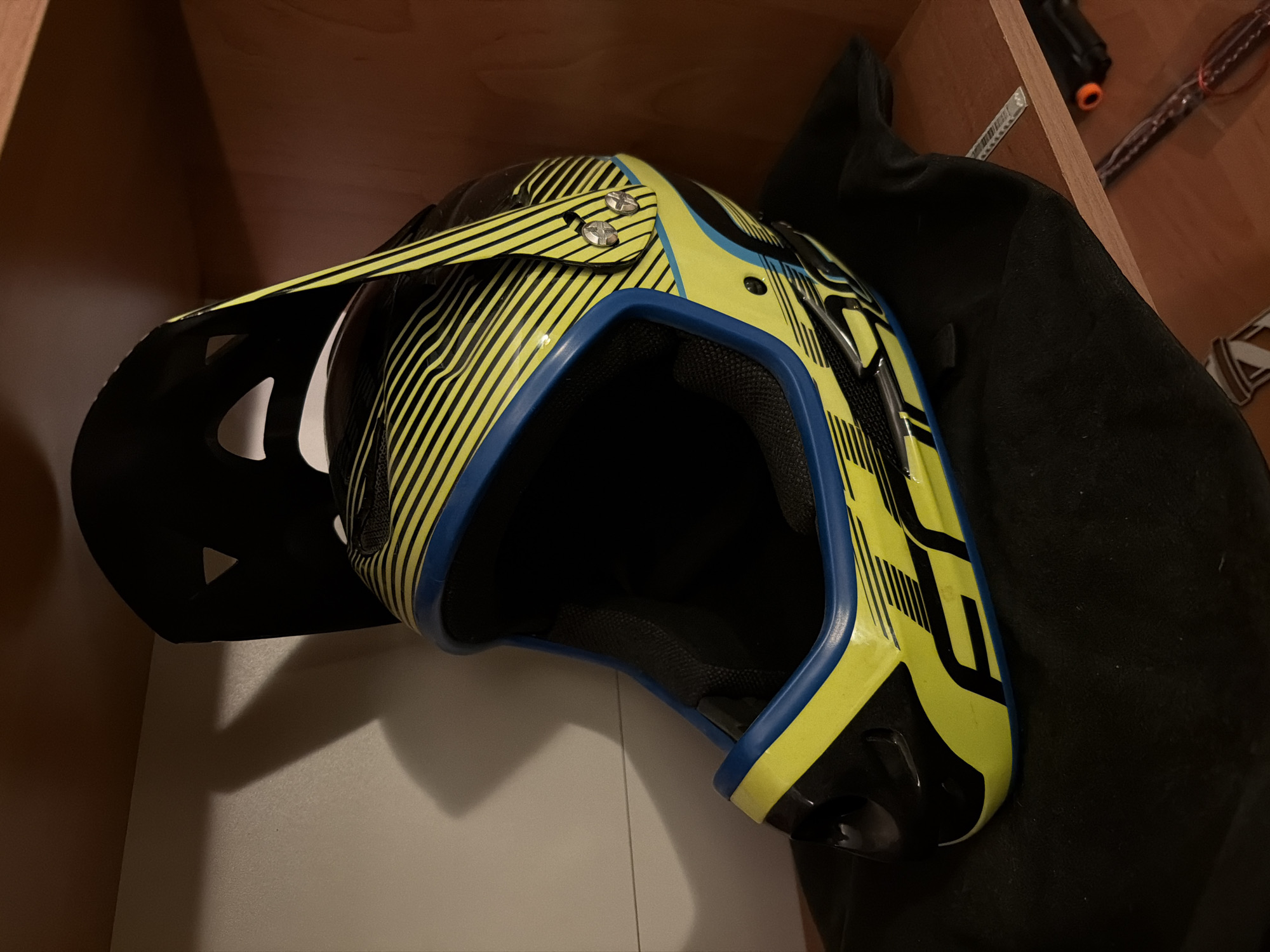 A yellow and blue mountain bike helmet is in a cupboard, positioned on its side with a black cloth behind it. The helmet has a black visor with triangular cutouts. The cupboard is made of light wood. In the background, a black object, measuring tape, and other items are partially visible. The location is unknown.