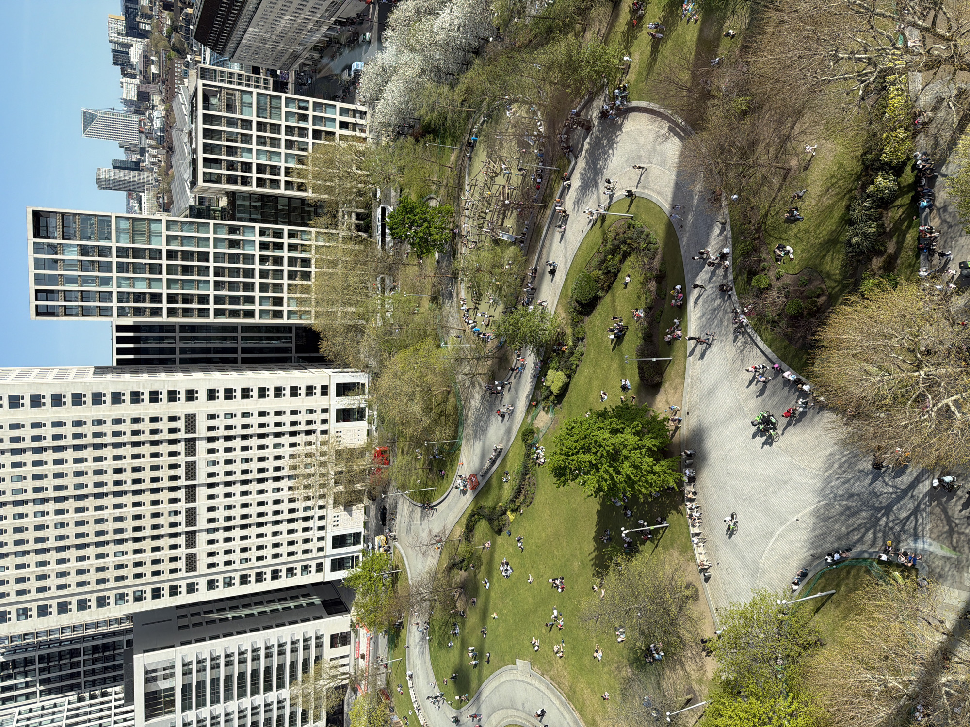 The image presents an aerial view of a park with buildings in the background. The park features meandering walkways populated by people walking, jogging, and cycling. Patches of green grass are occupied by individuals and small groups of people relaxing, picnicking, or sunbathing. Scattered trees provide shade and visual diversity. The architecture in the background consists of modern high-rise buildings with regular window patterns, suggesting an urban setting. Based on the landmarks and architectural style, this is a view of the area around Finsbury Avenue Square, located near Liverpool Street Station in the City of London, United Kingdom.