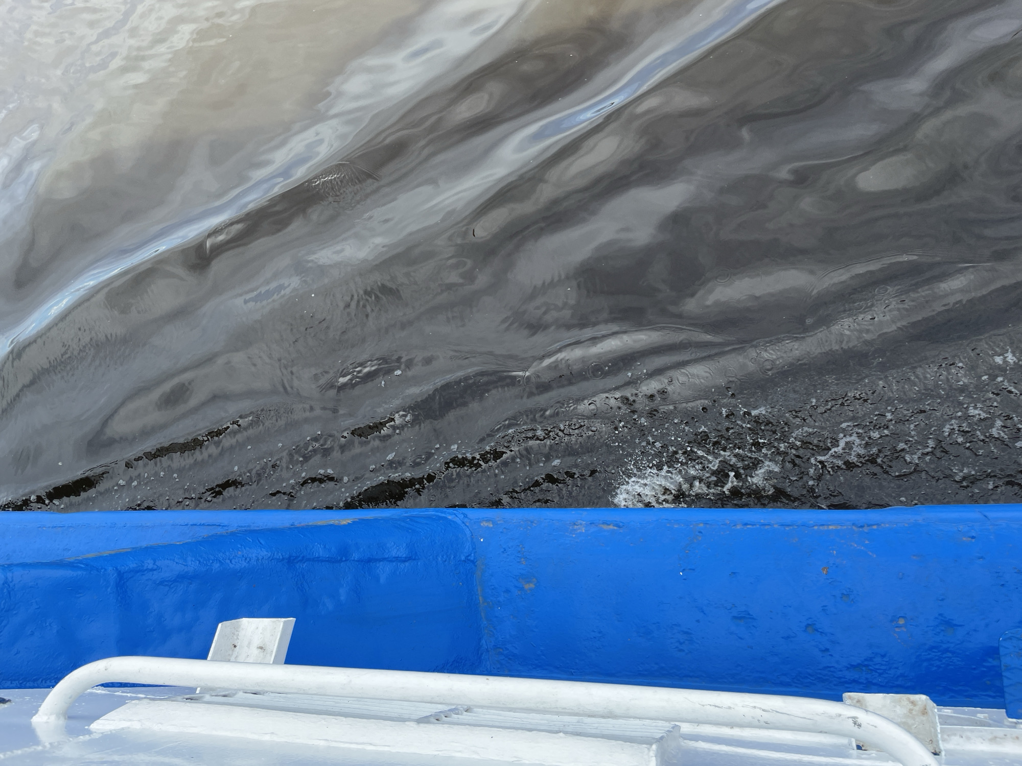 An overhead shot captures a section of water next to the blue hull of a boat, likely moving on a river in Perm, Russia. The upper two-thirds of the image are dominated by the water, which is a dark, swirling grey with lighter blue and silver reflections indicating ripples and an overcast sky. Patches of white foam and small splashes are visible where the water meets the boat's hull, suggesting movement. The lower third of the image shows the boat itself, with a distinct, vibrant blue hull exhibiting some wear and tear, including scuffs and small reddish-brown marks. Below the blue hull, a white deck or upper structure is visible, featuring a white metal railing or grab bar in the foreground. The overall lighting suggests an overcast day. No text is visible in the image.