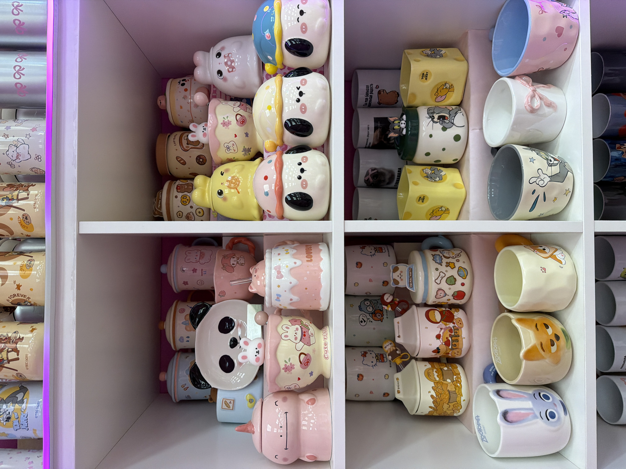The image shows a shelf display of novelty mugs, likely in a retail setting. The mugs are arranged in neat rows within the white shelving unit. The mugs vary in design, featuring cartoon characters like Tom and Jerry, animals like bunnies, dogs, and pandas, and themes such as "Zootopia". The shelves appear to be lit by a pink or purple light source, perhaps a neon strip or LED lighting, which casts a glow around the display. Some of the mugs have lids and sculpted handles. The surrounding texts contain the names of places in Russia (ПОПЛАКАЛА, АЛКАНАЛА, радость). The exact location of the scene in the city of Votkinsk, Russia, cannot be identified definitively from the image, but based on the text, the location must be a retail shop in Russia.