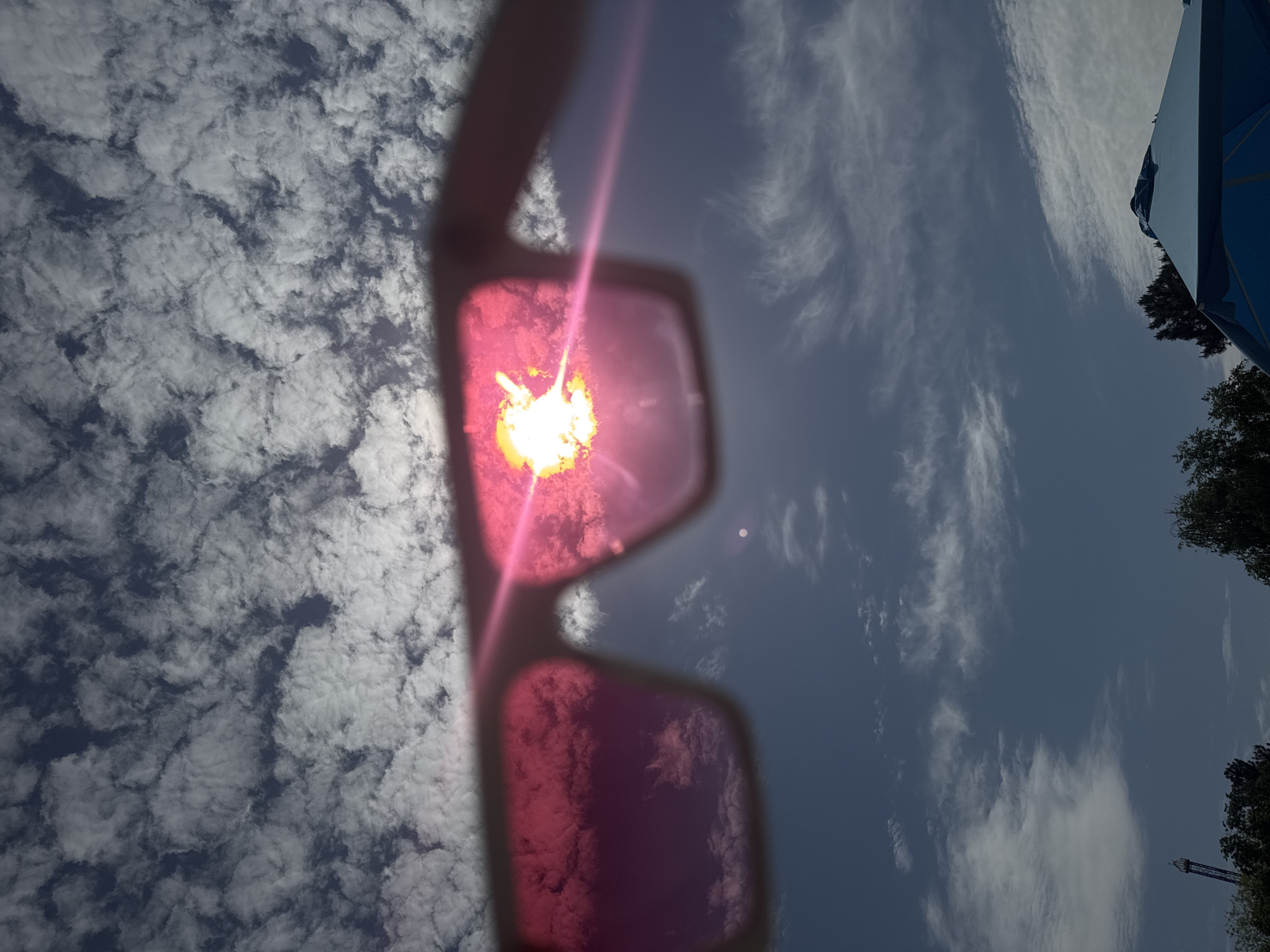 The image depicts a view of the sky through pink-tinted sunglasses. The right side of the sky exhibits cirrus clouds against a blue backdrop, while the left side shows cumulus clouds. The sun's glare is visible through the left lens of the glasses, appearing as a bright, radiant spot. A blue structure and trees are seen at the edge of the image. Based on the presence of trees and the absence of distinctive landmarks in the sky, the location cannot be pinpointed to a specific spot in Yevpatoriya, Ukraine from the given visual information.