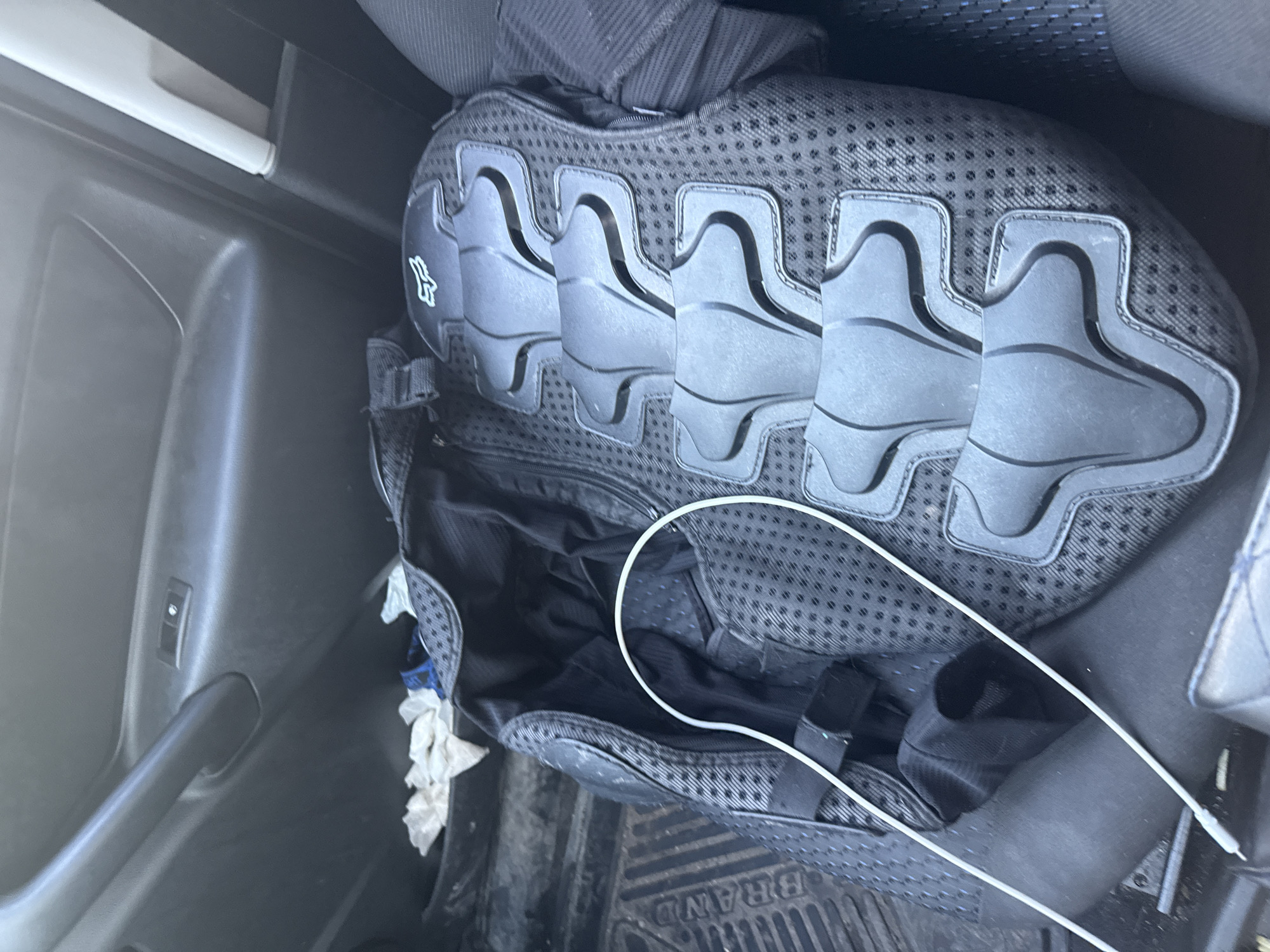 An overhead shot captures a black protective vest with segmented padding, resting inside what appears to be the passenger side of a vehicle. The vest is predominantly black, with a textured fabric base and distinct, curved grey protective panels running horizontally across its surface. A white logo, possibly a stylized fox head, is visible on the left side of the vest. The vest is partially unzipped, revealing a black interior with some crumpled white tissues peeking out. A thin white cable snakes across the vest and disappears into the opened compartment.
The vest is positioned on a black car floor mat, which has embossed text, partially visible, reading "BRAND". The surrounding environment is dark, suggesting the interior of a car. Visible on the car door to the left is a black door handle and a small silver switch or button. The lighting is dim, hinting at either a very early morning, late evening, or an overcast day, and the location cues are limited to the interior of a vehicle. There are no people or animals present in the image. The overall impression is one of stored gear, possibly for outdoor activities or sports, in a vehicle.