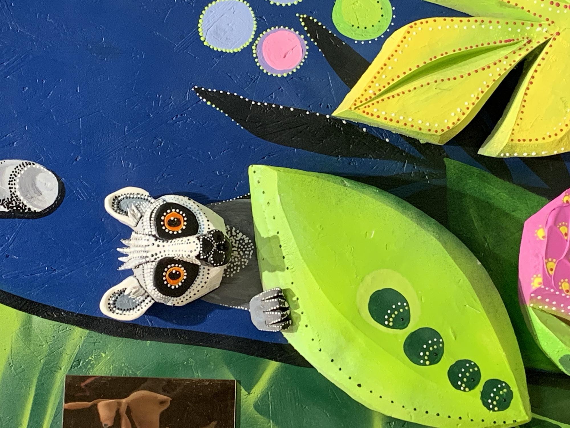 The image depicts an eye-level close-up of a colorful, possibly three-dimensional, artwork. The artwork features a lemur-like face with large, orange-ringed eyes and distinctive black and white patterned markings around the eyes and face. The lemur appears to be emerging from behind a large, green leaf with dark green circular spots. The lemur's paw is visible, gripping the edge of the leaf. To the right of the green leaf is a partially visible pink flower with yellow accents. Above, there is a yellow flower. There are also blue, pink, and green circles in the background, set against a textured blue backdrop. The background appears to be painted with visible brushstrokes. At the bottom is a small out of focus photo of an animal. The artwork is likely not located in the city of Stavropol, Russia.