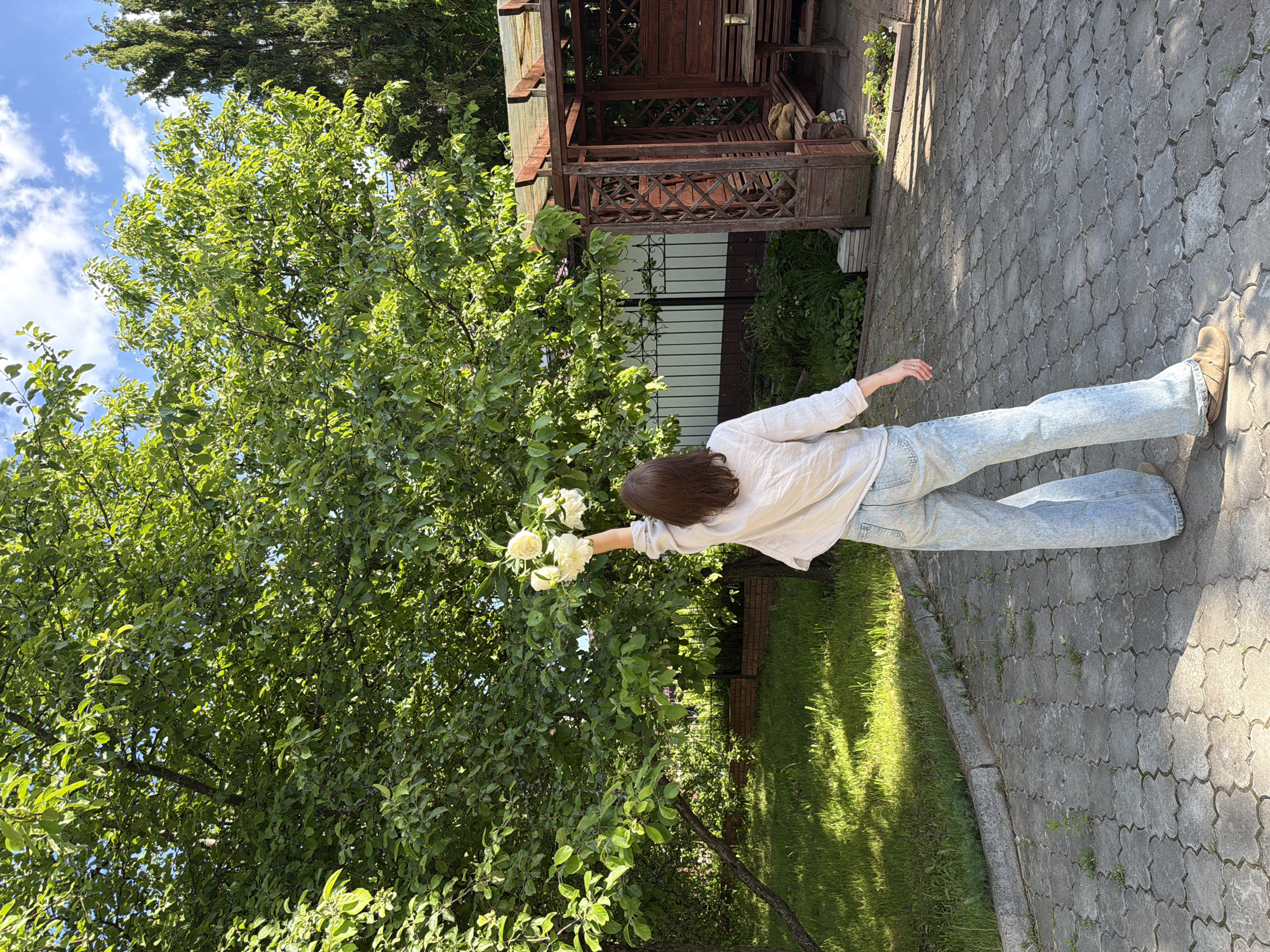 This image, captured in an outdoor residential setting in Прикамский, Russia, depicts a female individual standing with her back mostly to the camera. She has brown, medium-length hair and is dressed in a light-colored, long-sleeved shirt, light blue wide-leg jeans, and light brown slip-on shoes. Her right arm is extended upward, holding a bouquet of approximately 5-7 white or cream-colored flowers aloft, directed towards the dense green foliage of a large deciduous tree that dominates the left side and upper center of the frame.
The ground consists of grey, interlocking paver stones with a distinctive quatrefoil pattern, interspersed with small patches of green vegetation. To the left of the paved area, a section of vibrant green grass is visible, displaying distinct areas of sunlight and shadow. In the mid-ground on the right, a wooden gazebo with a dark brown stain, featuring lattice paneling and a partially translucent roof, is present. Inside the gazebo, a wooden bench and table are visible. A metal fence with decorative scrollwork stands in the background, backed by a light-colored wall and a darker lower section. Beyond the fence to the right, a tall, dark green coniferous tree rises. The sky above is blue with scattered white cumulus clouds, indicating fair weather and strong sunlight, which casts shadows across the scene.