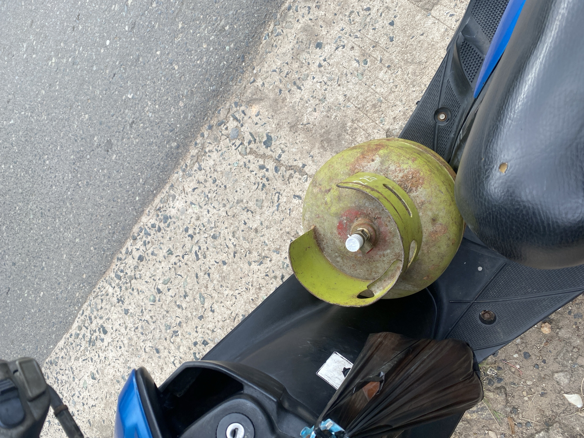 In Sungai Sipai, Indonesia, a gas cylinder sits on the floorboard of a scooter. The cylinder is primarily green with rust spots and a red top. A black plastic bag is tied to the scooter's handle with blue string. The scooter is parked next to a paved area with grey asphalt.