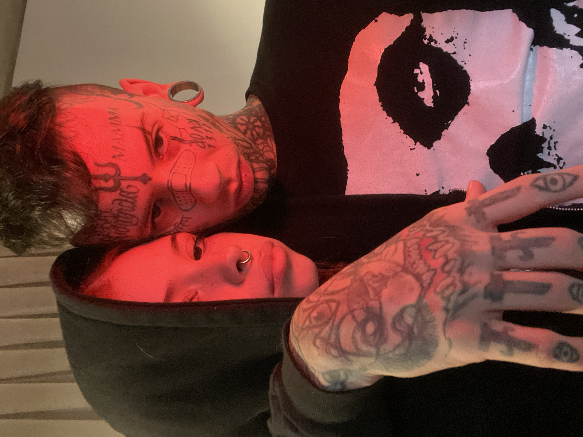 The image depicts two individuals indoors, illuminated by a red light source. The individual on top has extensive tattoos covering their face and neck. Visible tattoos include lettering, geometric shapes, and a drawing of a bandage. They have an ear gauge. They are wearing a black shirt with a white skull-like design. The individual below is wearing a black hooded garment and has a nose ring. Their right arm is visible with tattooed designs including eyes and other symbols. No specific details indicate a location in Buenos Aires, Argentina.
