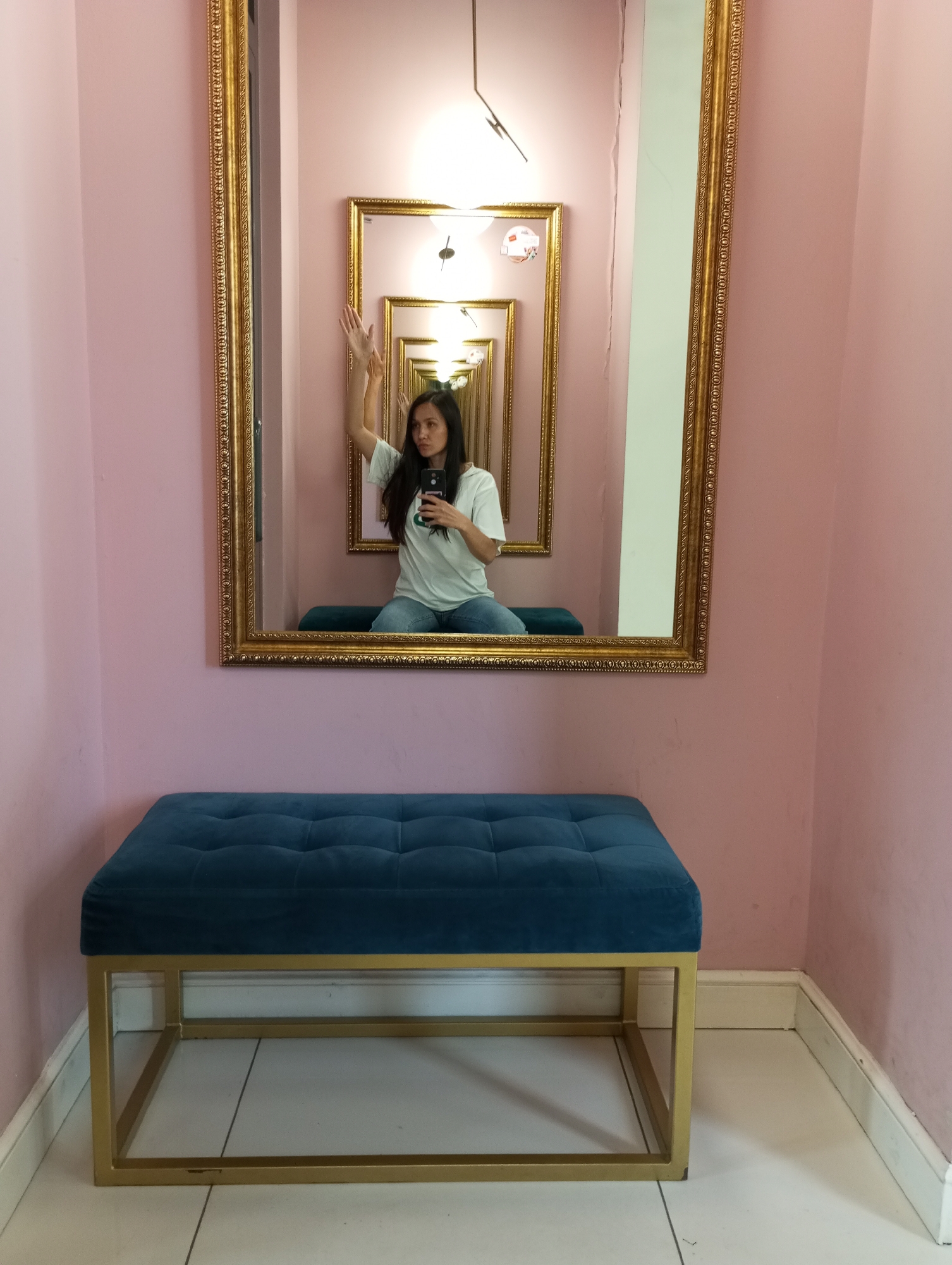 A medium, eye-level shot shows a woman in a mirror sitting on a blue tufted ottoman bench with a gold frame. The walls in the room are pink, and the tile floor is white. The woman in the mirror is wearing a t-shirt and jeans and holding a phone in her left hand while raising her right hand. The mirror is a large, ornate gold frame, and inside the mirror is a reflection of the woman in the mirror, creating a seemingly endless corridor of mirrors. The bench is in front of the mirror, and the room is well-lit. The location is in Izhevsk, Russia.