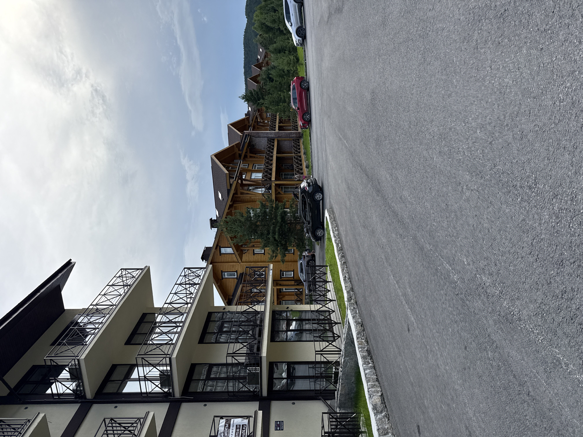 The image shows a modern multi-story building with balconies and large windows on the left, and several wooden-style apartment buildings with balconies in the background on the right. A paved road or parking lot occupies the foreground. Several cars are parked on the right side of the road. The buildings appear to be set in a wooded, hilly area with a cloudy sky. Signs are visible on the left building. The architecture suggests a resort or residential complex. The location is identified as Syrostan, Russia.