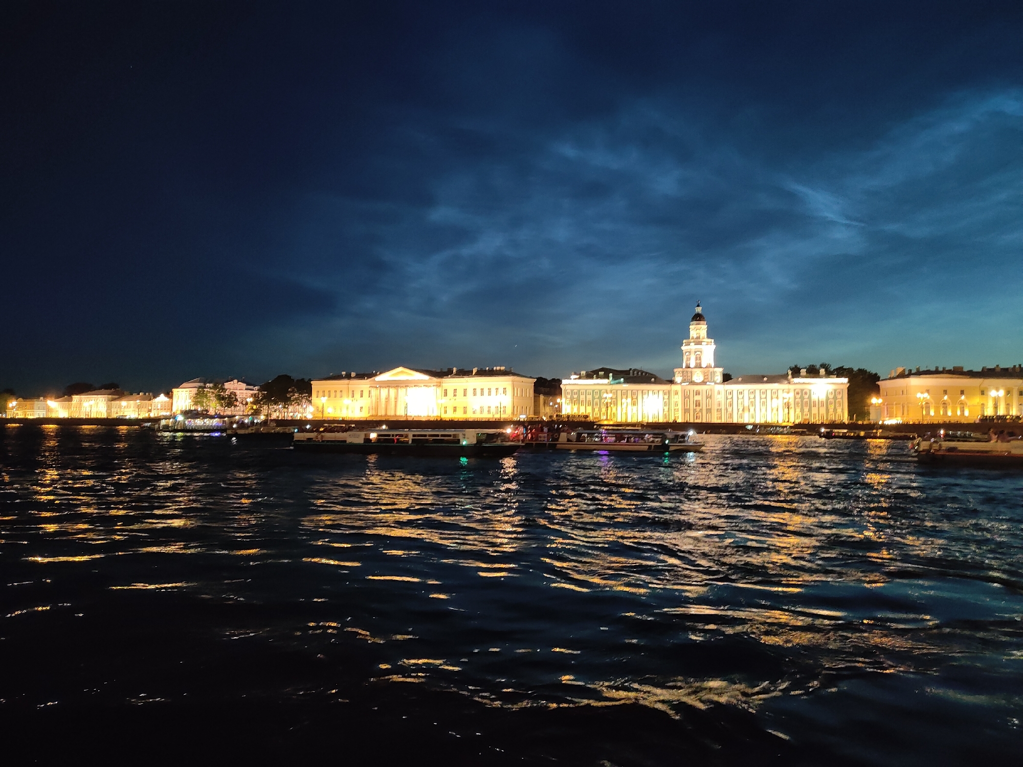 The image depicts a nighttime scene in Saint Petersburg, Russia, likely along the Neva River. In the foreground, the dark, rippling water reflects the warm, yellow lights of the buildings on the opposite bank. Several tour boats with illuminated windows are visible on the water, some carrying passengers. Across the river, a long, ornate building complex stretches along the embankment. This building is brightly lit, showcasing its classical architecture with numerous windows and detailing. To the right, a prominent tower with a clock and dome rises above the main structure, illuminated by spotlights. The sky above is a deep, dark blue with wispy, luminous clouds that appear to be noctilucent clouds, a phenomenon often visible in Saint Petersburg during the summer months. The overall atmosphere is serene and picturesque, capturing the architectural beauty of the city at night.
