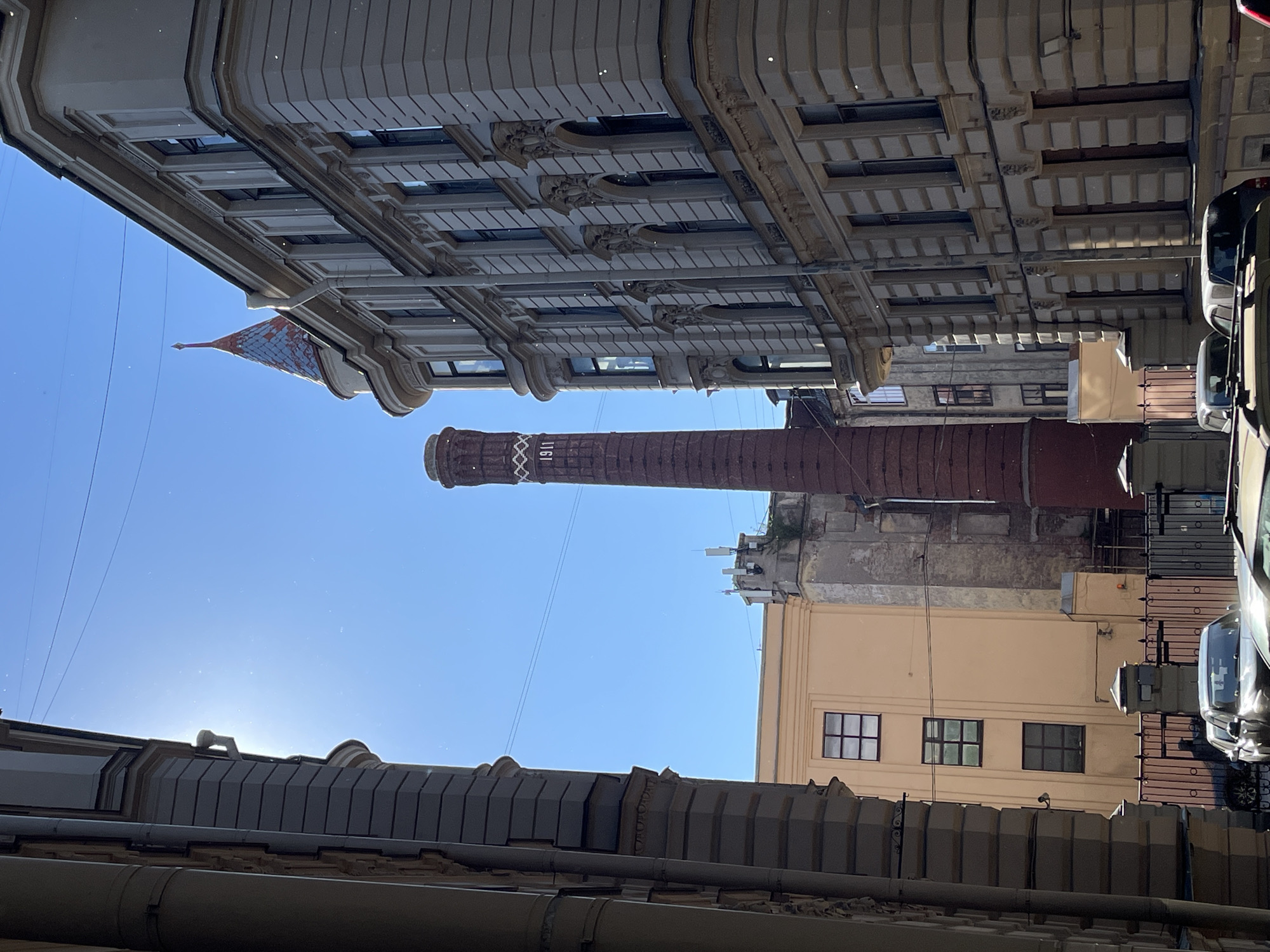 The image depicts a narrow urban street in Saint Petersburg, Russia, with buildings on either side and a clear blue sky. The perspective is from below, looking upwards, and the entire image is rotated 90 degrees counterclockwise, meaning the top of the image is on the left, and the bottom is on the right.
The dominant subjects are the architectural elements of the buildings. On the left, a building with a light-colored facade features ornate detailing along its upper floors, including decorative moldings and a series of regularly spaced windows. At the top of this building, a distinctive pointed roof or spire with a patterned surface is visible. In the center of the frame, a tall, reddish-brown industrial chimney rises against the sky. White lettering is visible on the chimney, reading "1911". To the right, another building with a pale yellow facade has a simpler design with several dark-framed windows. The foreground and bottom of the image are filled with the dark, textured roofs and structures of buildings. Cars are visible parked along the right side of the street, suggesting a populated area.
The scene appears to be taking place during the daytime, with bright sunlight casting shadows and illuminating the sky. The weather seems clear and sunny. The presence of the industrial chimney and the historical architecture hints at the industrial past of the city. No people are clearly visible in the frame. The overall impression is of an old, possibly historic, urban environment.