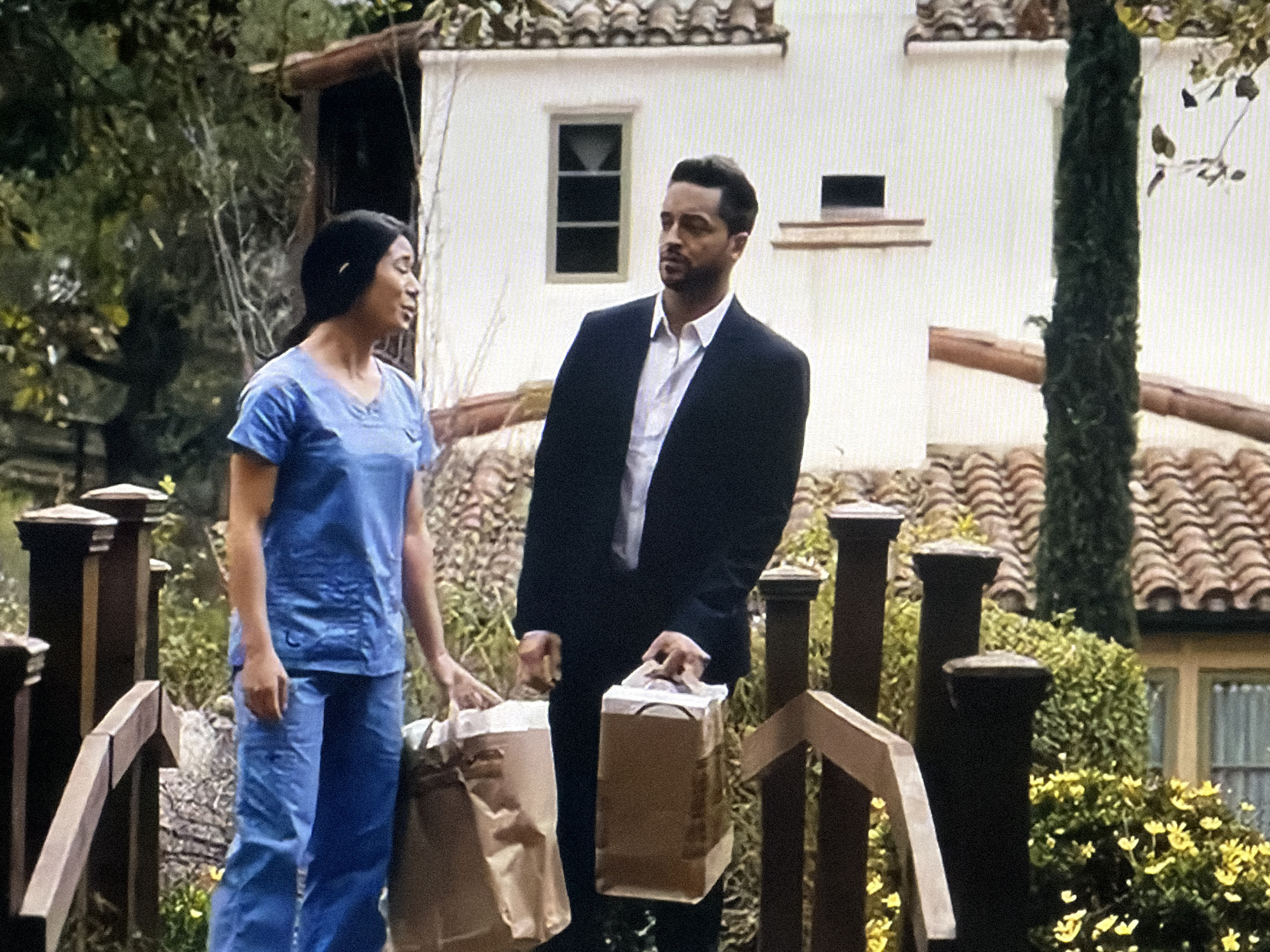 Two individuals stand outdoors, appearing to exchange or deliver items in broad daylight. On the left, a woman with dark hair, wearing light blue medical scrubs, holds a large brown paper bag. Her head is turned towards the man, and her mouth is slightly open, suggesting she is speaking. On the right, a man with dark hair and a neatly trimmed beard, dressed in a black suit jacket over a white collared shirt, holds a smaller brown paper bag. He stands facing generally forward but with his gaze directed slightly to the right of the frame, away from the woman.
They are positioned in front of a white building with a reddish-brown tiled roof, characteristic of Mediterranean or Spanish-style architecture. A small, dark rectangular window is visible on the building's facade. Lush green trees, bushes, and smaller plants with small yellow flowers frame the scene, indicating a warm and verdant environment. A dark wooden railing or fence structure is prominent in the foreground, partially obscuring the lower half of the figures and adding depth to the composition. The sky is bright, and the overall impression is of a clear day. No identifiable text is visible on the paper bags or elsewhere in the image.