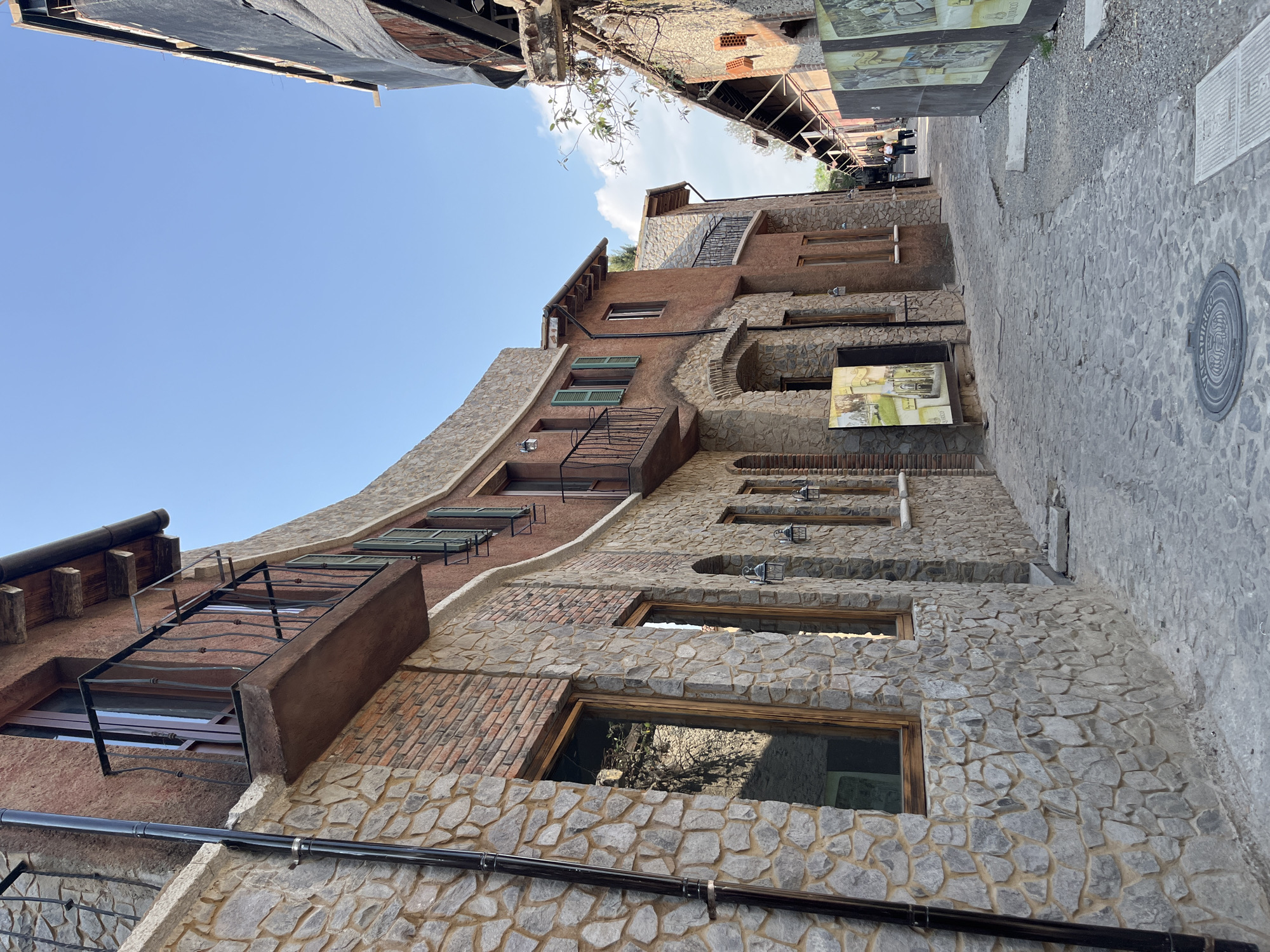 The image depicts an upward view of an unusual building with mixed materials in what appears to be a narrow street. The facade of the building consists of a combination of gray stone, red brick, and painted red-brown sections. There are various windows and architectural features like archways and external staircases. Overhead, glimpses of the sky and roofs of other buildings are visible, suggesting a densely built environment. The paving of the street appears to be stone-cobbled, indicating a historic or traditional setting. The location is Domingo Arenas, Mexico. There are no people or actions visible in the immediate scene. A series of signs and displays are mounted on the wall along the right side of the street.