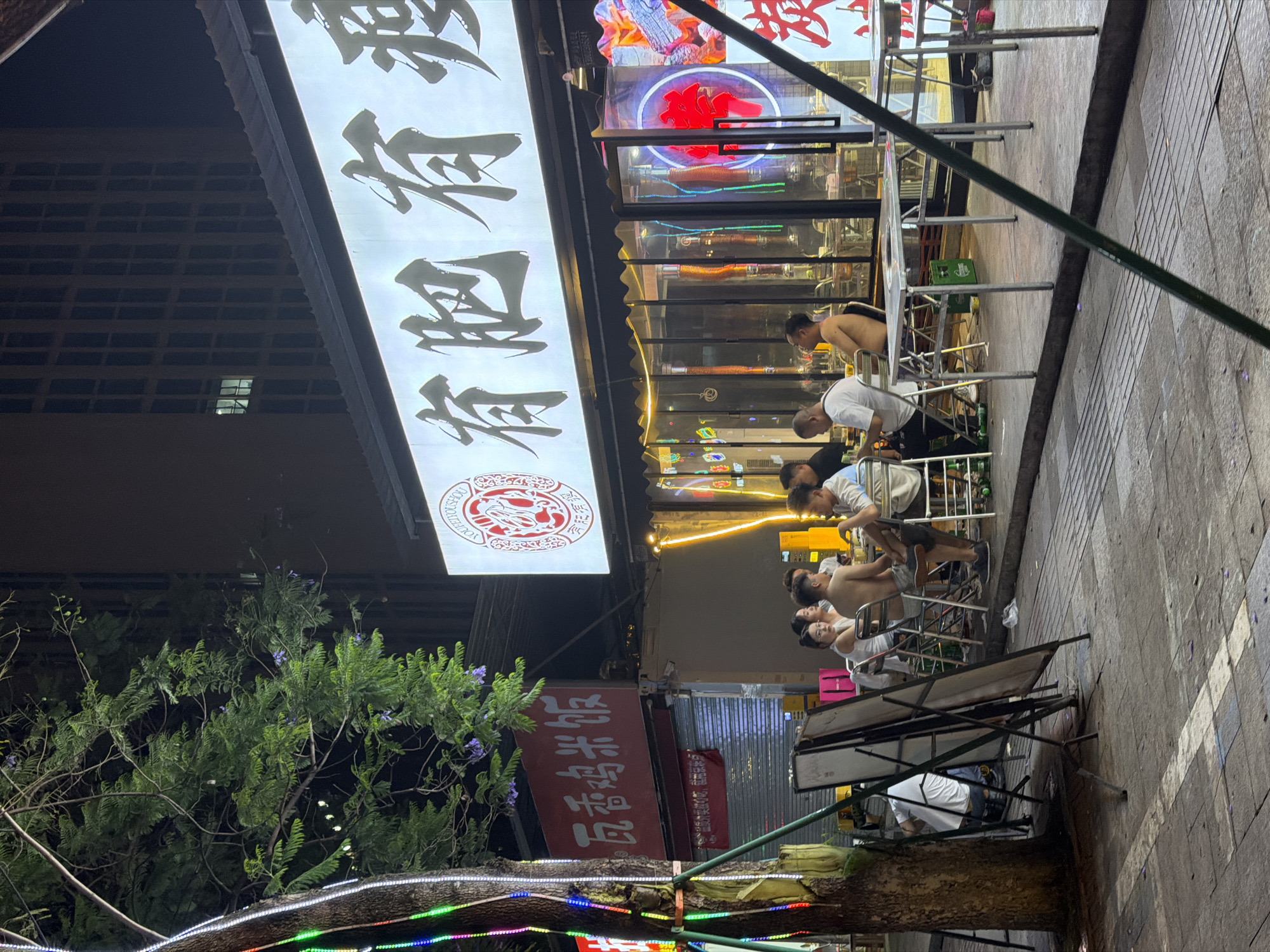 The image shows an outdoor dining scene at night in Chongqing High-tech Zone, China. Several people are sitting at metal tables and chairs eating. A sign with Chinese characters hangs above the restaurant, with another sign featuring Chinese characters and the word "YOUHOUTOUCHENG" below it. The ground is paved with rectangular stones. A tree with colorful lights wrapped around its trunk is visible on the left side of the image. A multi-story building is partially visible in the upper left.
