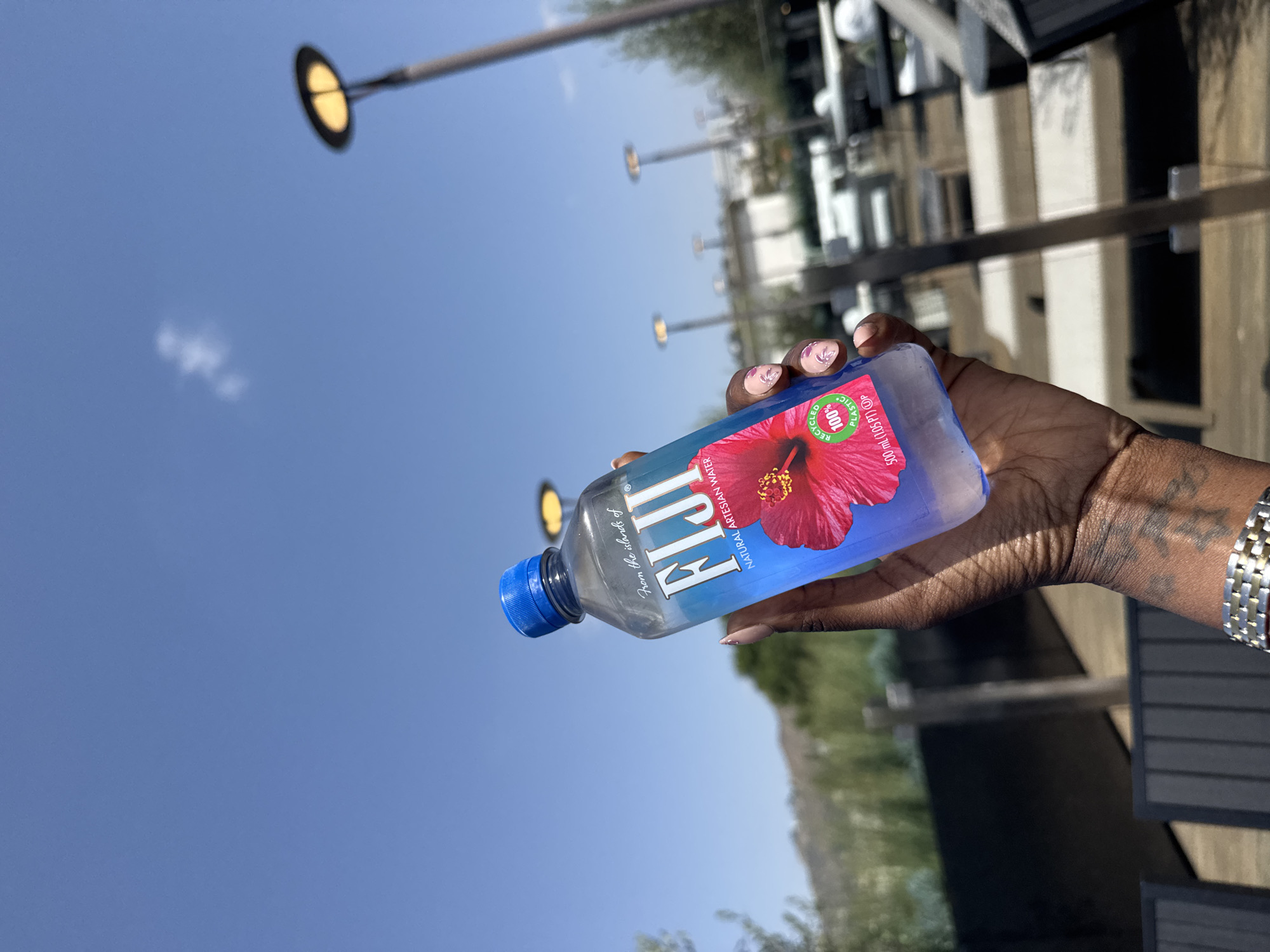 Here is a detailed description of the image:
A hand holds a bottle of Fiji water with a hibiscus design. The bottle is blue with a blue cap. The background shows an outdoor area with a clear blue sky and multiple street lights. There are buildings and greenery in the background as well. The photograph appears to be taken during the daytime. The location could be within the city of Los Angeles, United States of America, given the typical urban and clear sky landscape. The person has manicured nails. The person wears a bracelet. The person has tattoos on the hand. The writing on the bottle reads: From the Islands of Fiji, Natural Artesian Water, Recycled 100% Plastic.