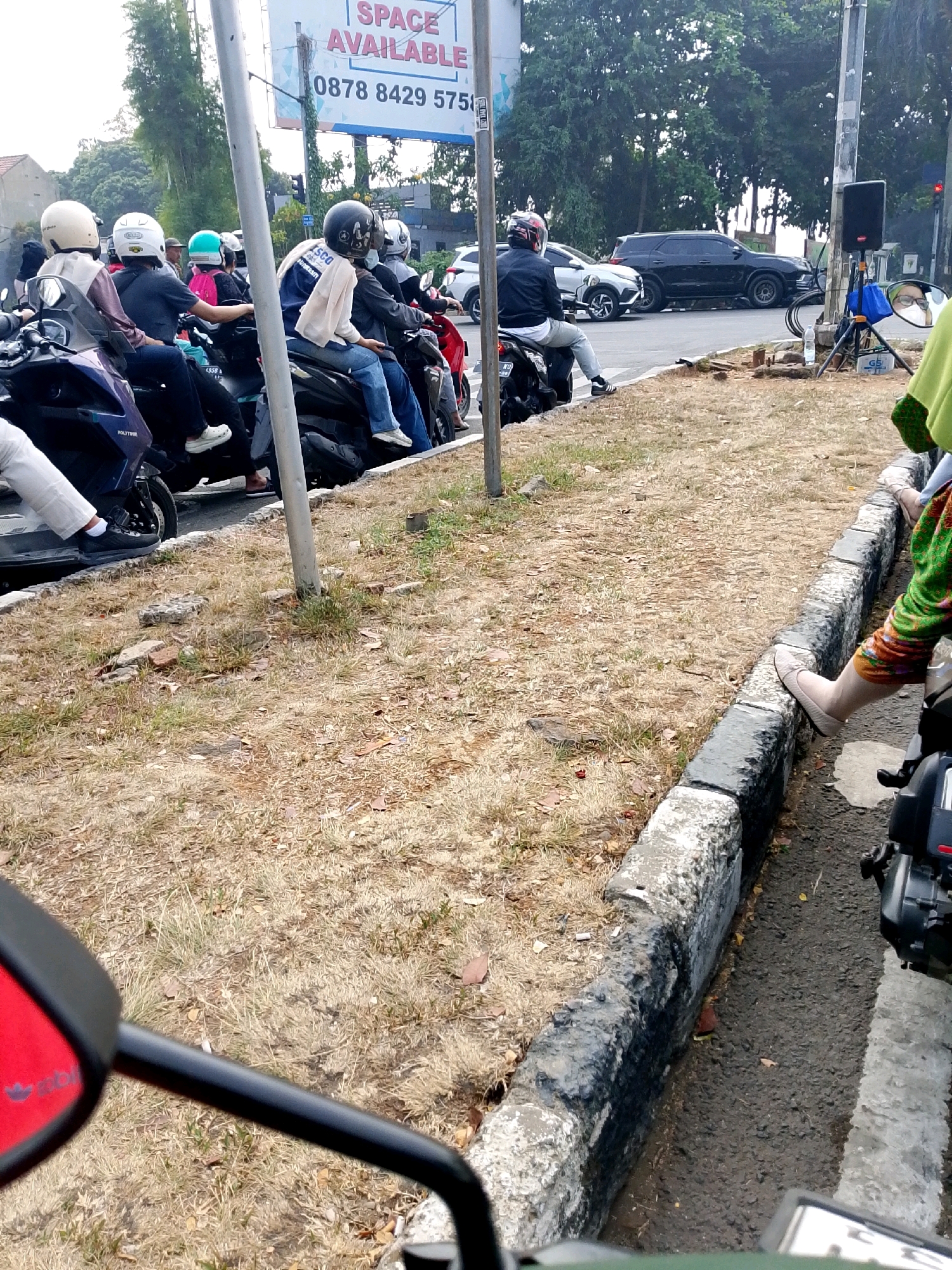 The image captures a traffic scene in Cibinong, Indonesia, likely near a main road given the presence of several vehicles and a billboard. Several motorbikes are lined up, with riders wearing helmets. The riders appear to be waiting, possibly at a traffic signal or in congestion. One billboard in the background advertises "SPACE AVAILABLE" and a phone number, 0878 8429 5751. A black SUV and other vehicles are visible on the road. The environment features dry, sparse grass along the roadside and a concrete curb. The overall impression is a typical urban street scene in Indonesia during daylight.