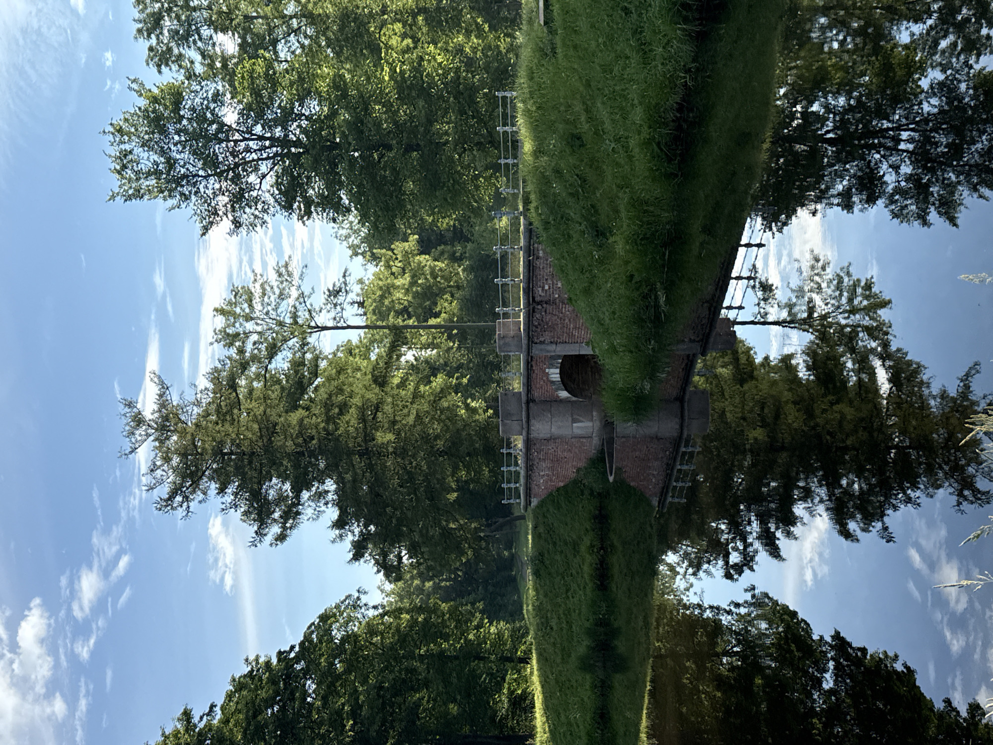 The image depicts a scenic view of a park in the city of Pushkin, Russia, likely during daytime. A brick arch bridge spans a calm body of water in the foreground, with its reflection clearly visible on the water's surface. The bridge is flanked by lush green grass and mature trees on both sides, creating a tranquil, natural environment. The sky above is a clear blue, dotted with white clouds, suggesting fair weather. The overall scene is one of serene parkland, characteristic of a well-maintained garden or estate.