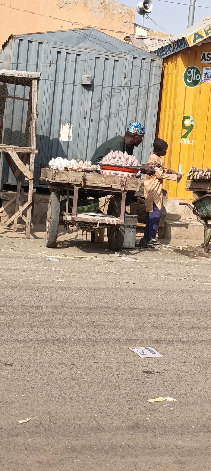 The scene depicts a street-level commercial area in Kaura Goje, Nigeria, under daylight conditions. Two individuals are present: an adult male and a child male.
The adult male, with dark skin and wearing a dark green long-sleeved shirt and a patterned blue and white cap, stands behind a two-wheeled wooden vendor cart. This cart displays multiple piles of light-colored, knobby produce, consistent with ginger or garlic, and a red-rimmed white enamel basin containing a reddish, granular substance.
The child male, with dark skin and dressed in a light tan tunic and dark blue trousers with green sandals, stands to the right of the main vendor cart. He holds a silver-bladed knife in his right hand, positioned towards dark, segmented stalks (likely sugarcane) arranged on a smaller, green metal cart.
In the background, two repurposed shipping containers serve as commercial structures. To the left, a weathered gray corrugated metal container exhibits partial white graffiti. To the right, a bright yellow corrugated metal container features prominent "glo" and "9 MOBILE" telecommunication logos, with the text "ABBAN UTAMA" visible above. Behind these structures, a faded orange-brown building wall with peeling plaster is visible. A white horn speaker is mounted on a pole above the yellow container.
The ground is a gray asphalt surface, sparsely scattered with small pieces of debris, including organic matter and a white paper wrapper with blue text in the foreground.