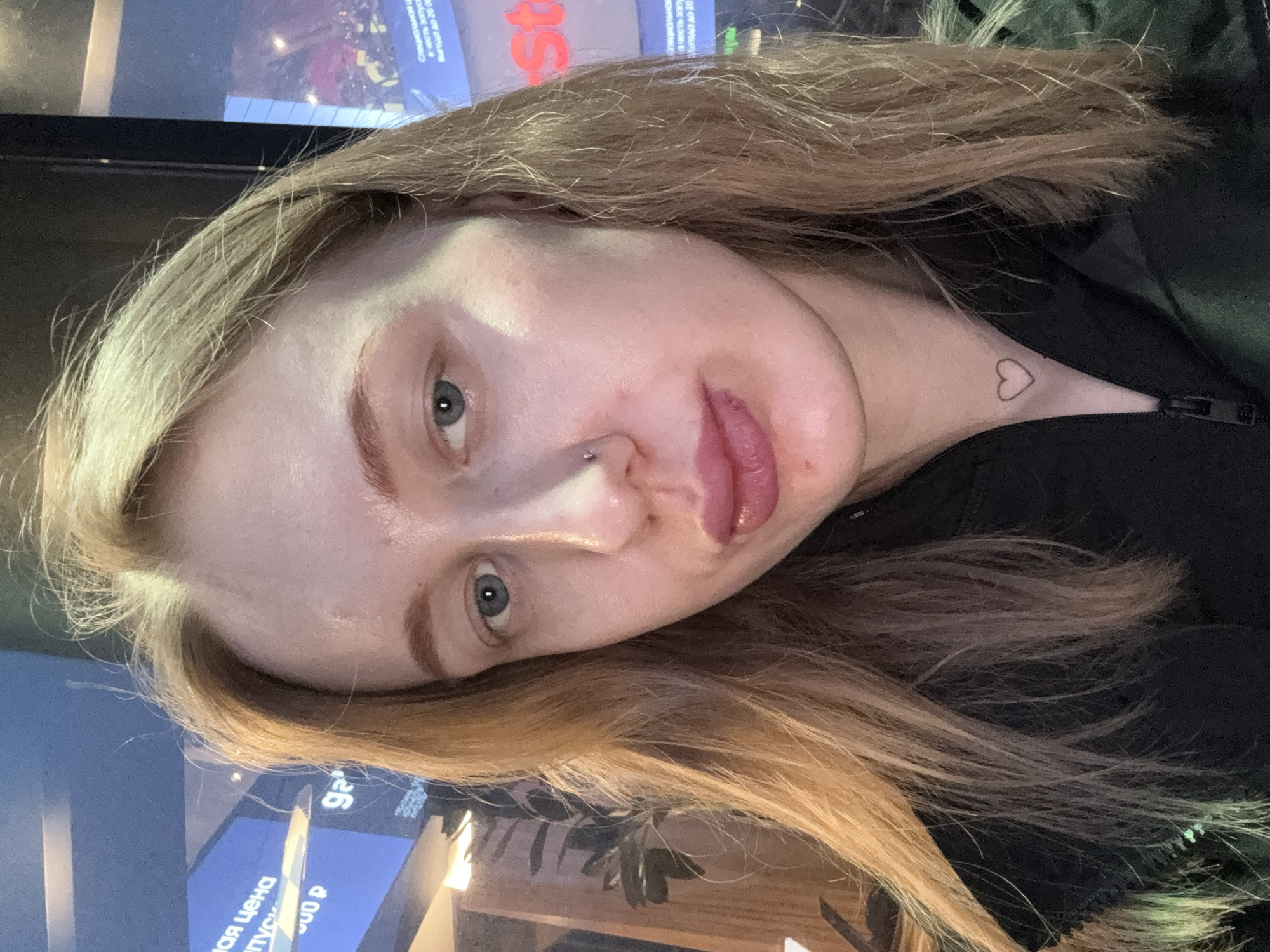 A close-up, rotated selfie of a fair-skinned young woman with light brown/blonde hair. She has a nose piercing and a heart tattoo on her neck. The background is slightly blurred and includes parts of screens displaying text in Cyrillic script. The background text includes “мая цена” and “Выгода до 20 00”. The image appears to have been taken indoors, possibly in a public space. There is no specific indication of Ol’ginskaya, Russia.