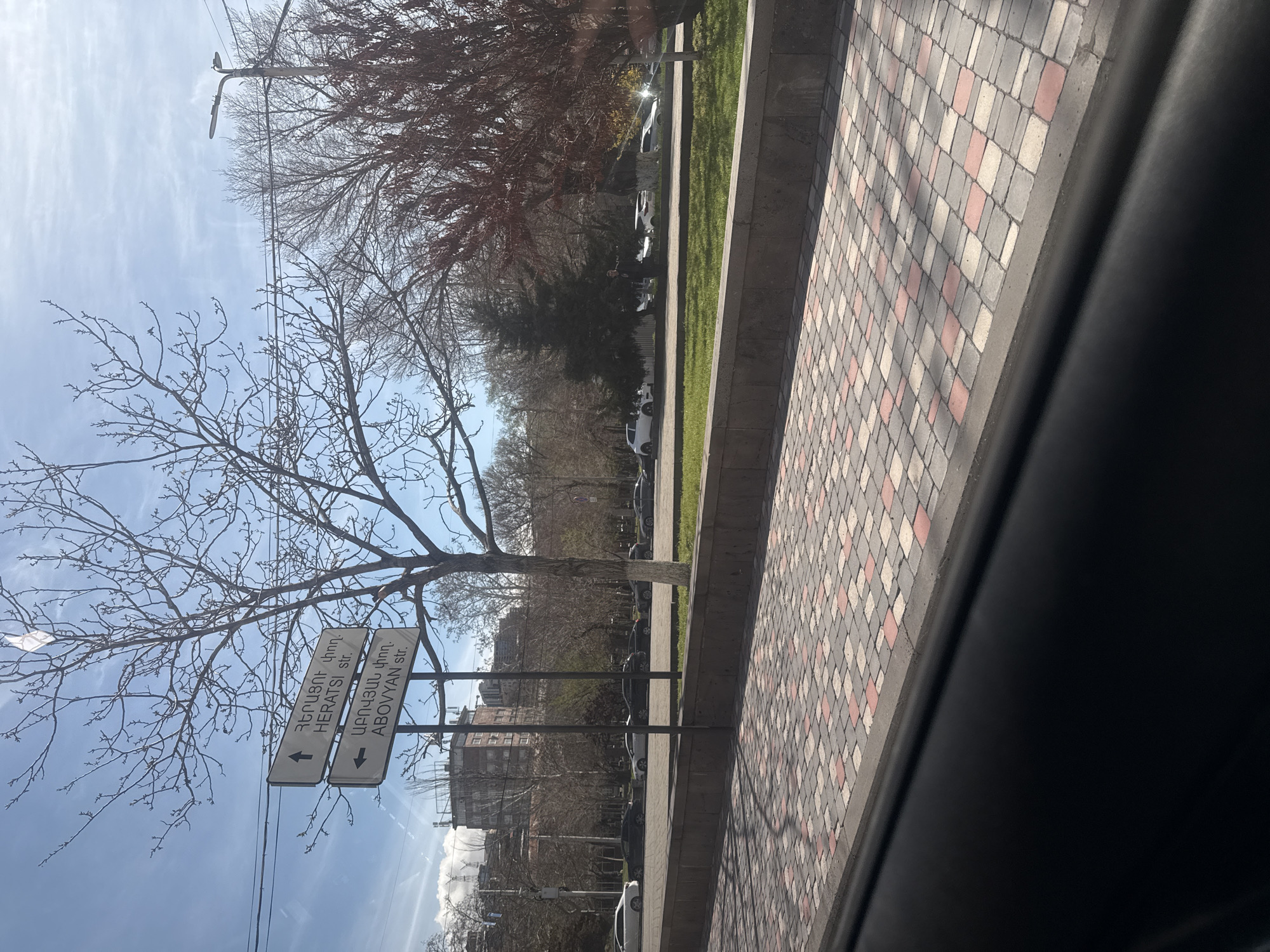 The image depicts a street scene in Yerevan, Armenia. A street sign indicates directions to Heratsi Street (left) and Abovyan Street (right). The foreground features a paved sidewalk with a pattern of grey, white, and red bricks, partially obscured by a dark object (likely the edge of a vehicle window). Beyond the sidewalk, there is a green strip of grass followed by parked cars. Bare trees are present, silhouetted against a light blue sky with thin clouds. Buildings can be seen in the background. A lamppost is visible amidst the trees. The image seems to be taken from inside a vehicle.