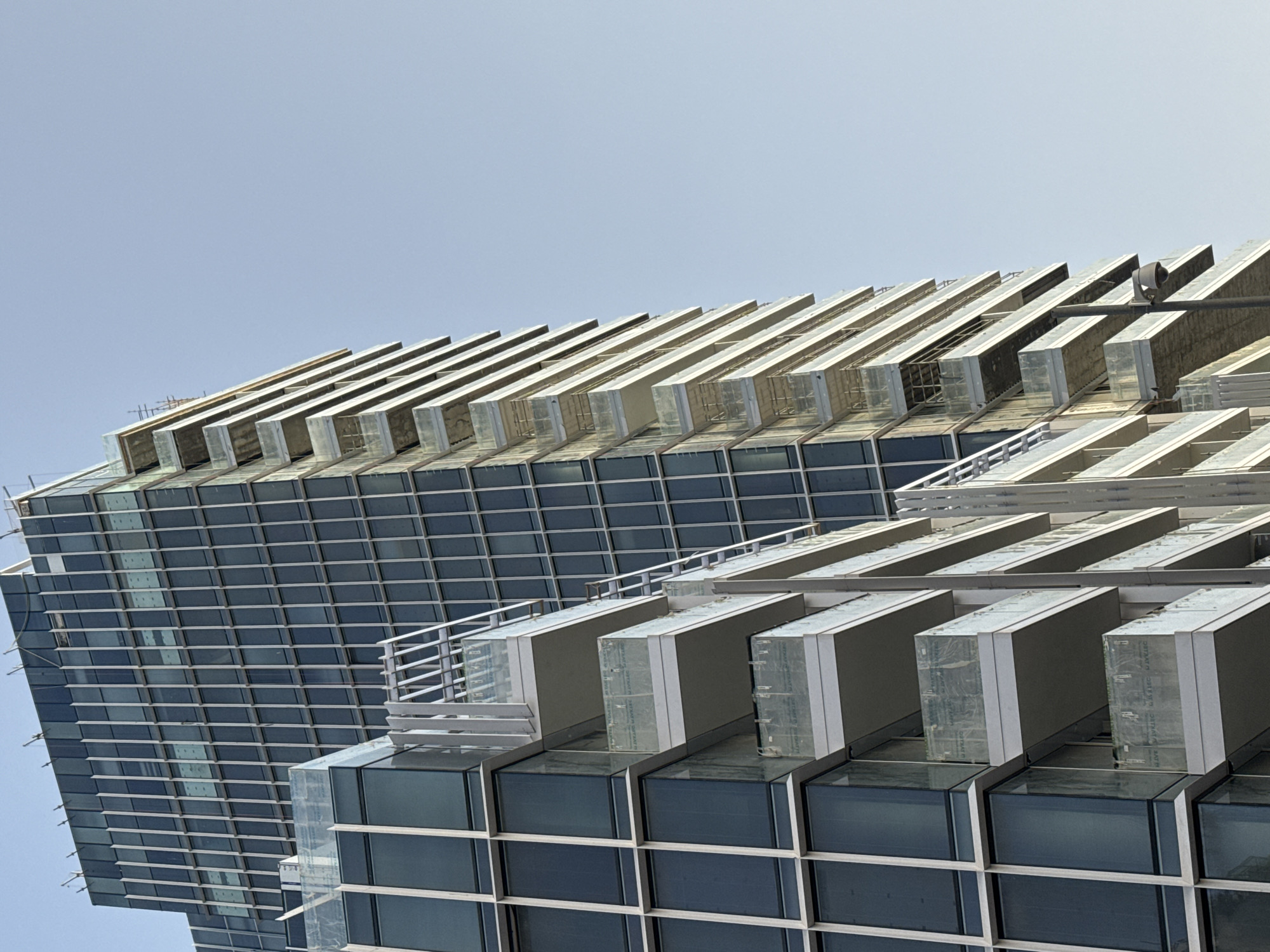 The image shows an upward-facing view of two modern buildings with a blue sky as a backdrop. The buildings are characterized by a grid-like pattern of dark windows with light-colored frames. The upper sections of the buildings feature a protruding, horizontal element that creates a visually layered effect. The architecture suggests a contemporary style, with an emphasis on geometric shapes and reflective surfaces. The image is taken in Tel-Aviv, Israel.
