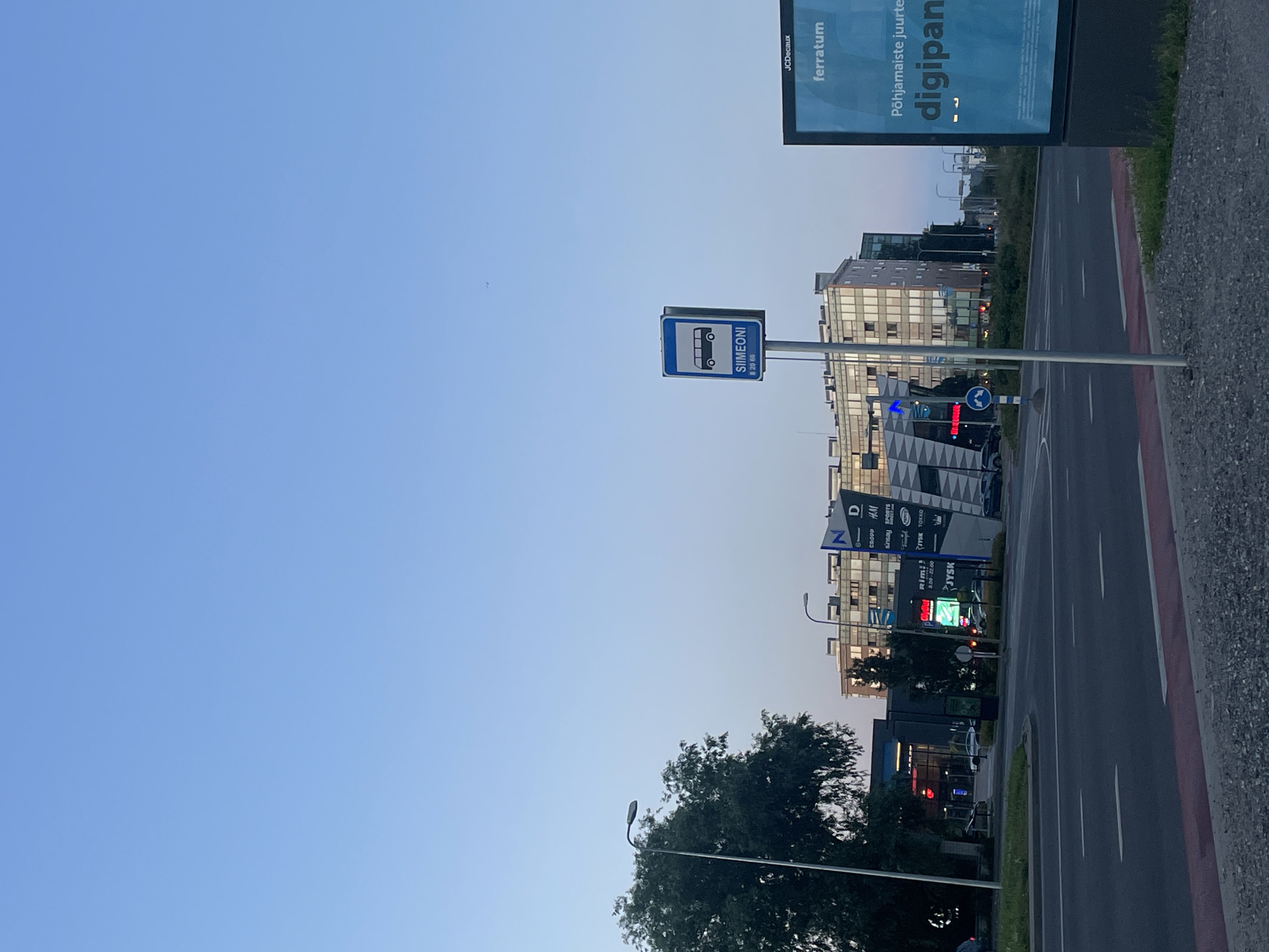 The image shows a street scene in Tallinn, Estonia, at dusk. In the foreground, a gray asphalt road with white lane markings is visible. To the right, a large blue digital billboard displays text in Estonian, including "Põhjamaiste juurtele digipani" and "JCDecaux ferratum". In the mid-ground, a bus stop sign with a bus icon and the text "SIIMEONI 8 20 66" is mounted on a pole. Behind the bus stop, a modern multi-story building with a geometric facade and numerous windows stands. Several storefronts are visible on the ground floor of this building, including signs for "N", "D", "CROPP", "H&M", "SINSAY", and "SPORTS DIRECT". Further to the left, another building with illuminated signage, possibly a supermarket or retail complex, is present. A large, leafy tree stands to the left of the street, and streetlights are visible throughout the scene. The sky is clear and blue, transitioning to a lighter hue near the horizon, indicating the end of daylight. There are no people or vehicles clearly depicted in the image.