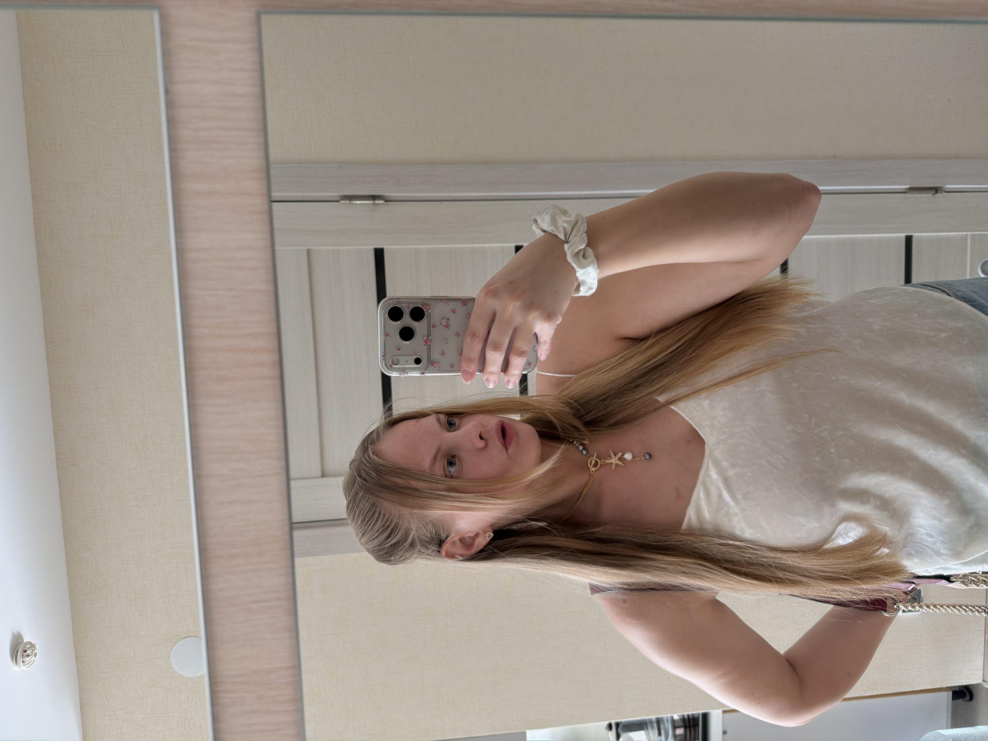 A woman is taking a selfie in front of a mirror. She has long blonde hair and is wearing a white top and a necklace. She is holding a smartphone with a floral case. A white scrunchie is on her wrist. The background appears to be an interior space with a door. The location in the city of Izhevsk, Russia is unknown.
