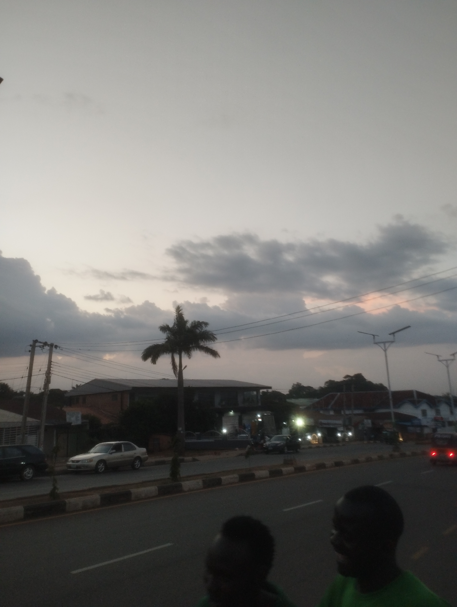 The image captures a street scene at dusk in Jos, Nigeria. The sky is overcast with dramatic, dark clouds tinged with the warm colors of sunset. A silhouetted palm tree stands prominently against the sky, with power lines and utility poles crisscrossing the frame.
On the street, several cars are visible. One silver sedan is parked by the roadside, while other vehicles are driving along the asphalt road. A row of buildings lines the opposite side of the street, some appearing to be commercial establishments with lights on, suggesting evening activity. Lampposts with modern designs are spaced along the sidewalk.
In the foreground, two individuals are partially visible, their faces turned away from the camera or obscured by shadow, with one person wearing a green shirt. Their presence suggests people are out and about in this urban environment. The overall mood is calm, with the fading light and cloudy sky creating a somewhat muted atmosphere. The scene depicts a typical evening on a busy street in a Nigerian city. No visible text is present in the image.