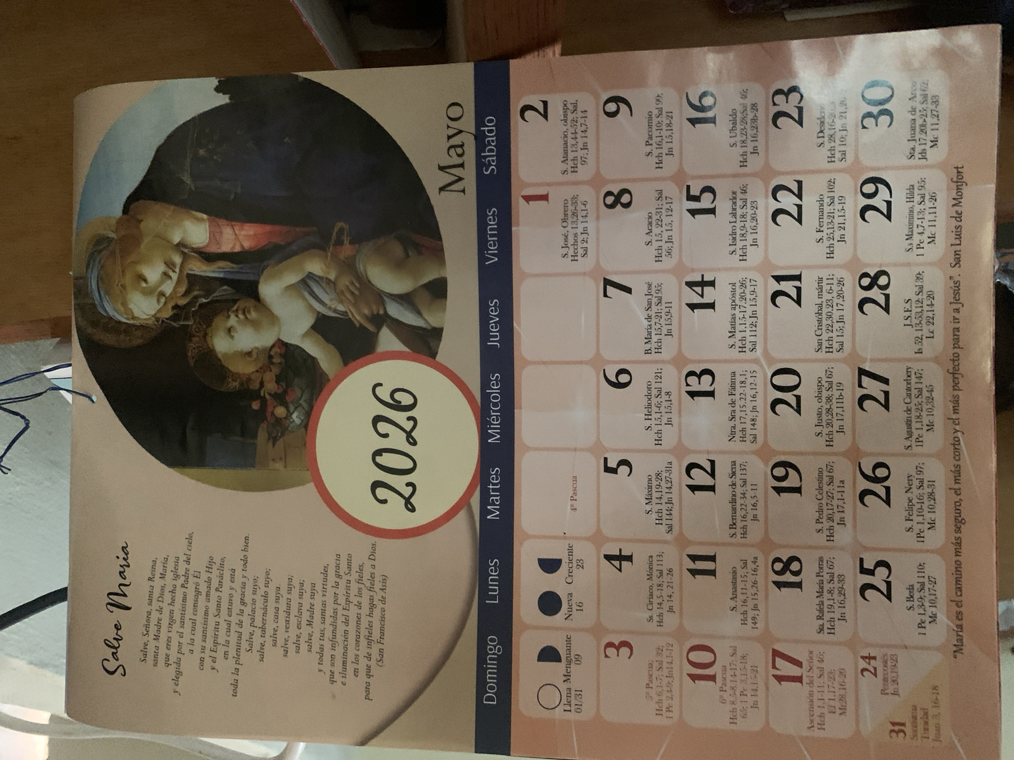 The image is a photograph of a calendar for May 2026. The calendar page features a depiction of the Virgin Mary and the Christ child. The calendar includes religious quotes in Spanish and the days of the month with associated religious figures and Bible verses. There is no location in the city of Santa Ana, Venezuela, visible in the image.