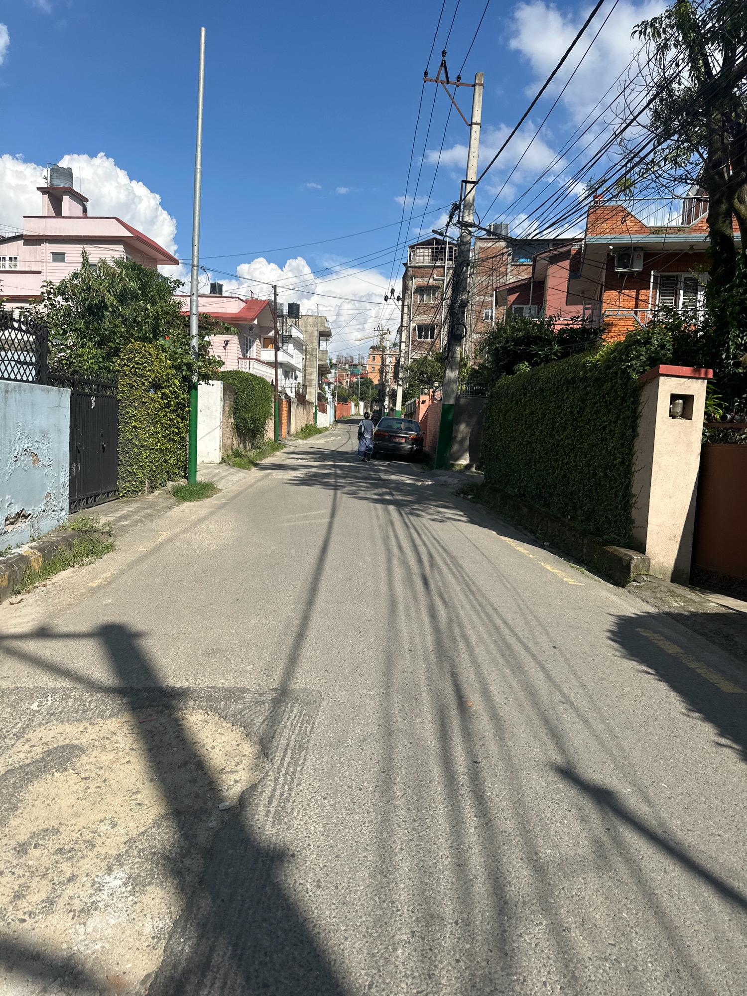 The image shows a residential street in Lalitpur, Nepal, on a sunny day with scattered clouds. The road is paved and runs between rows of houses and walls. On the left side of the street, there is a light blue wall with peeling paint and a black metal gate, partially obscured by lush green foliage. Further down, a pink house with a red roof is visible. On the right side, a tall, thin metal pole stands near a cluster of pink houses. Further along the right, a red brick house with a dark roof is adorned with greenery. A dense green hedge lines the right side of the street for a significant portion of its length, leading to a cream-colored pillar with a light fixture. Power lines crisscross overhead, casting long shadows on the asphalt. A dark-colored car is parked on the right side of the road, and a person in a light-colored outfit is walking away from the camera in the distance on the right. The overall impression is of a quiet, well-maintained urban neighborhood.
