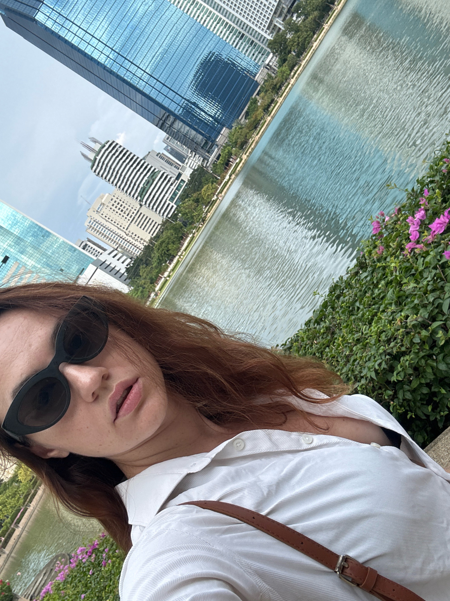 A woman with reddish-brown hair and sunglasses is taking a selfie in a park in Bangkok, Thailand. She is wearing a white button-down shirt. In the background, there is a body of water with buildings and trees along the bank. The buildings are modern, with glass facades. There are pink flowers in the foreground.